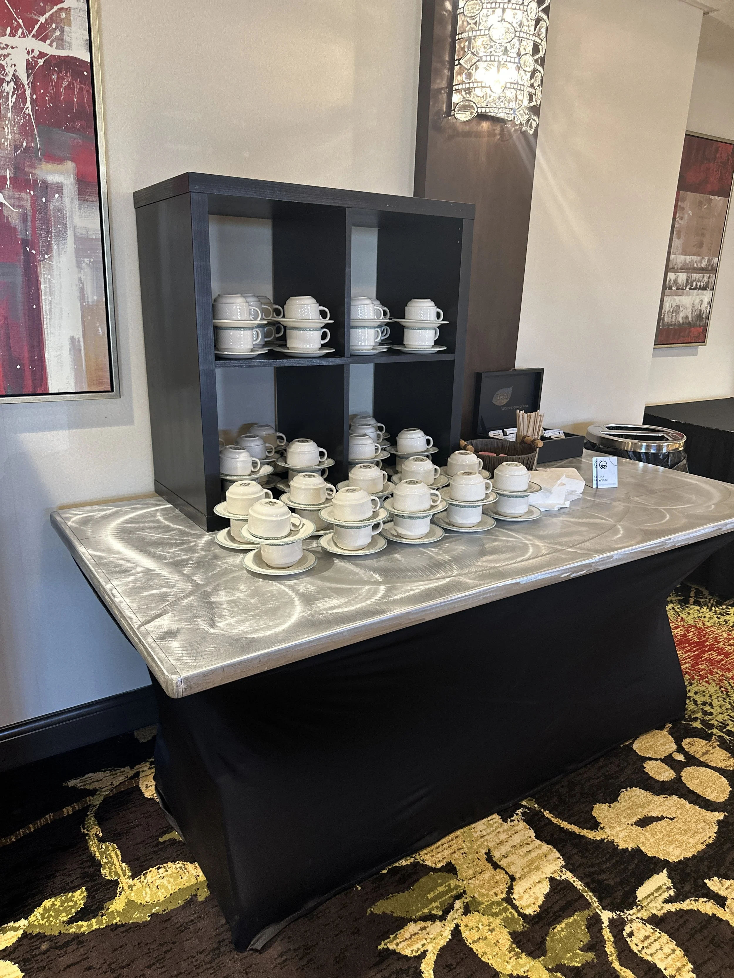 A table with stacked white coffee cups and saucers arranged in a display, with a black shelf holding additional cups in a hotel or conference setting.