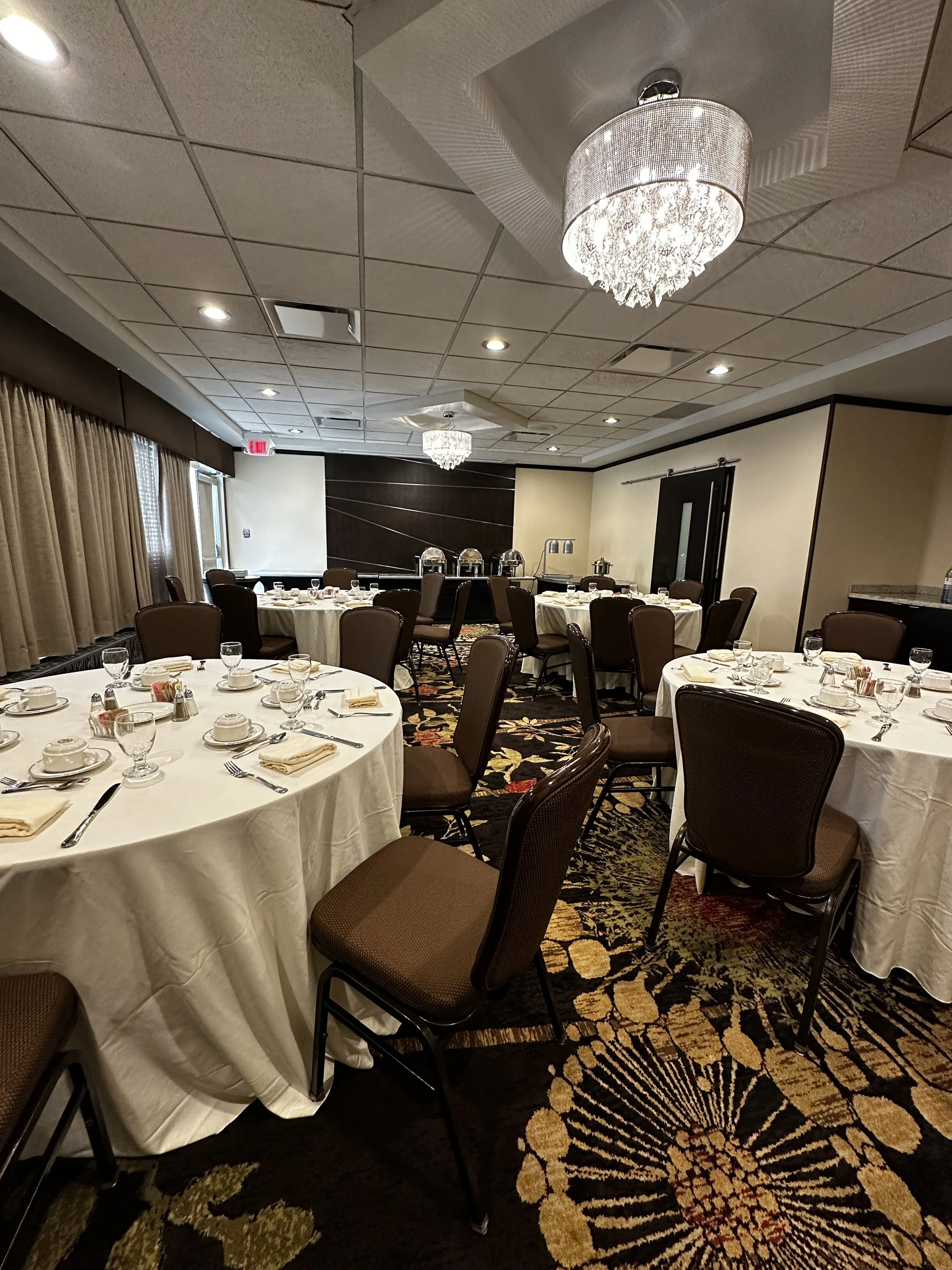 A banquet room set up for a formal event with round tables covered in white tablecloths, each set with place settings including plates, glasses, silverware, and napkins. Multiple brown chairs surround the tables. There are chandeliers hanging from the ceiling, and a buffet area with covered chafing dishes at the back of the room. The room has beige walls, dark accents, and a large window with curtains.