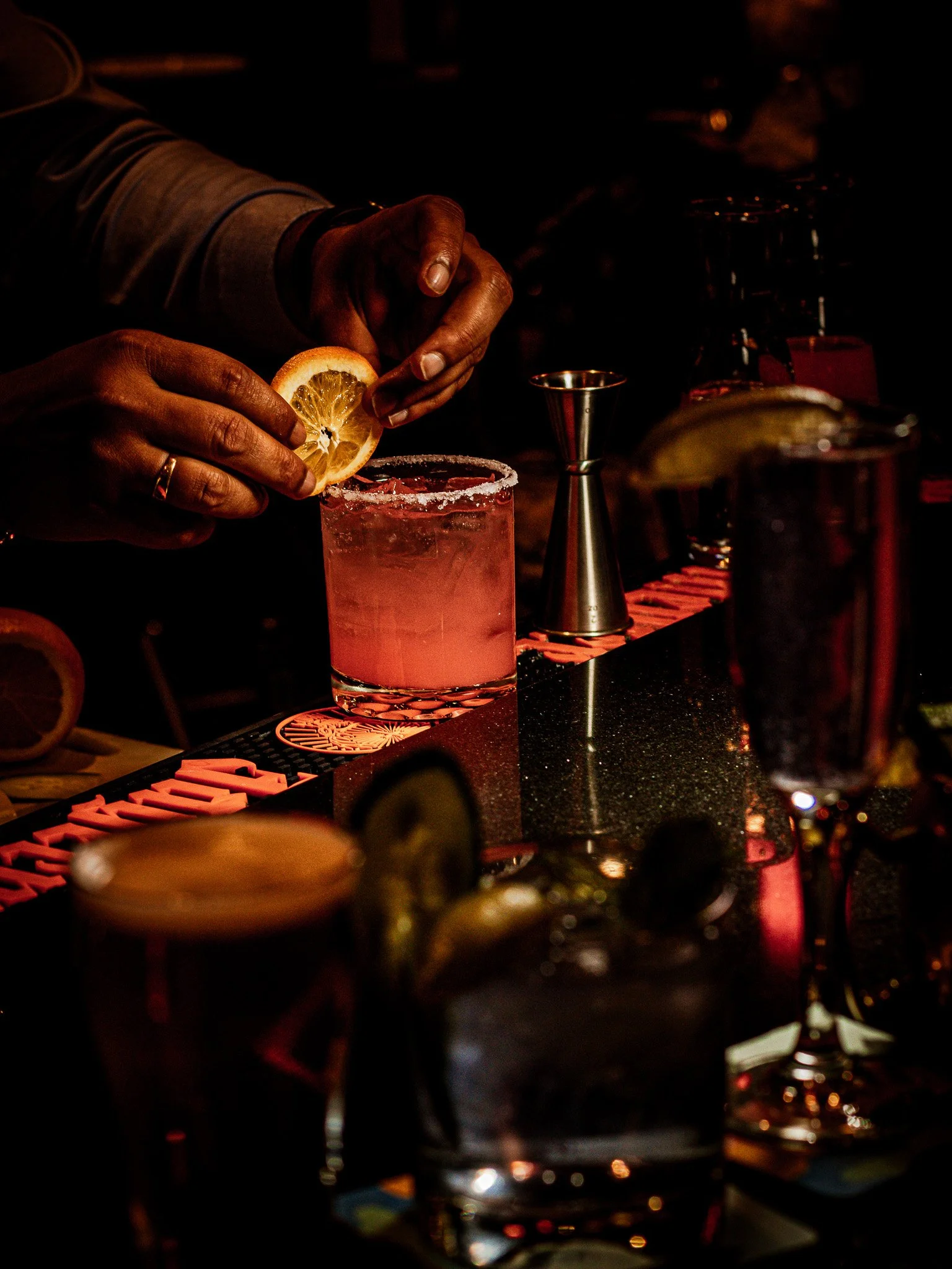 Person garnishing a pink cocktail with a lemon wedge in a dimly lit bar.