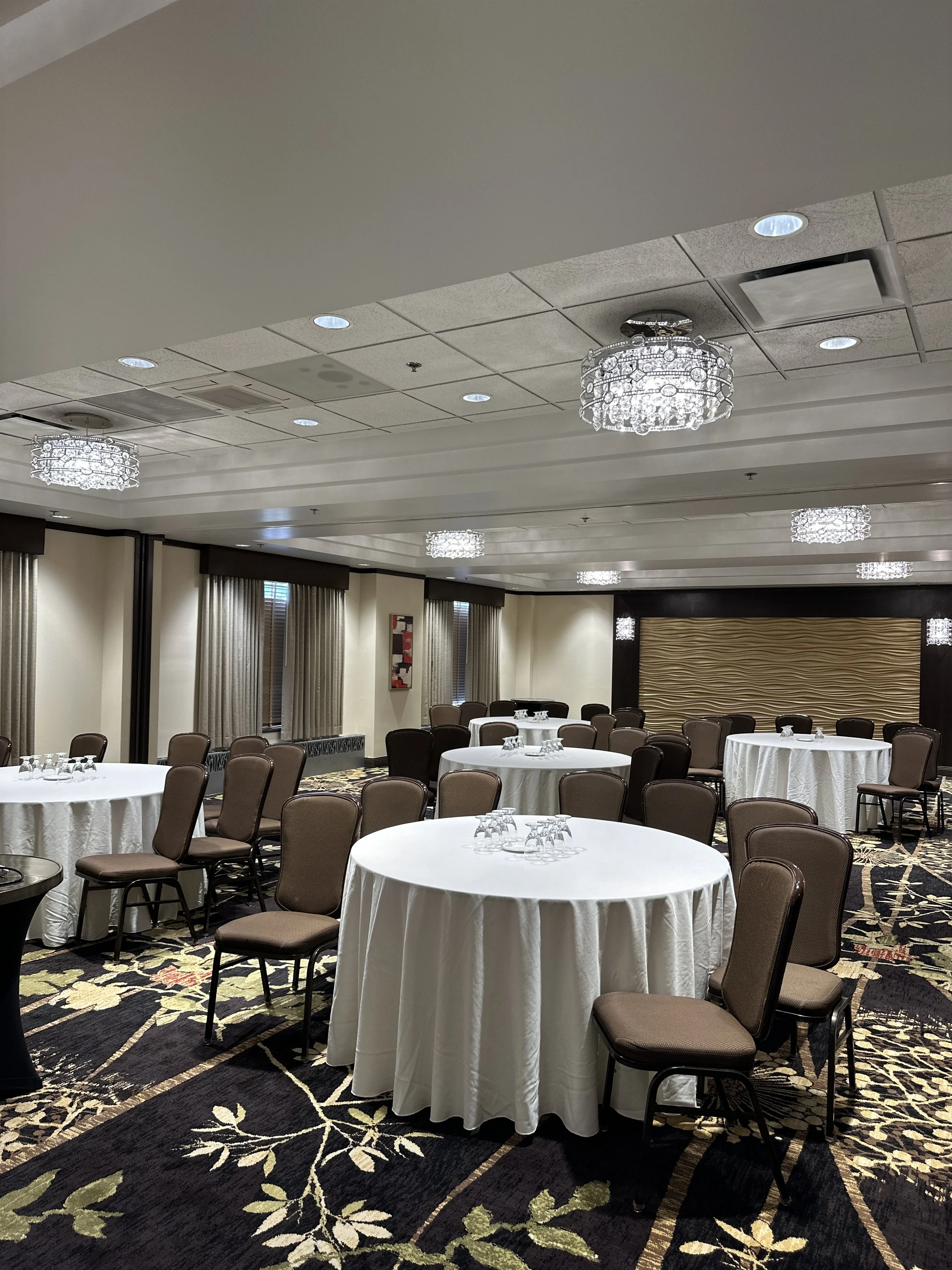 Banquet room with round tables draped in white tablecloths, surrounded by brown chairs, and decorated with glassware. The room has a carpeted floor with floral patterns, large windows with curtains, and ceiling chandeliers.