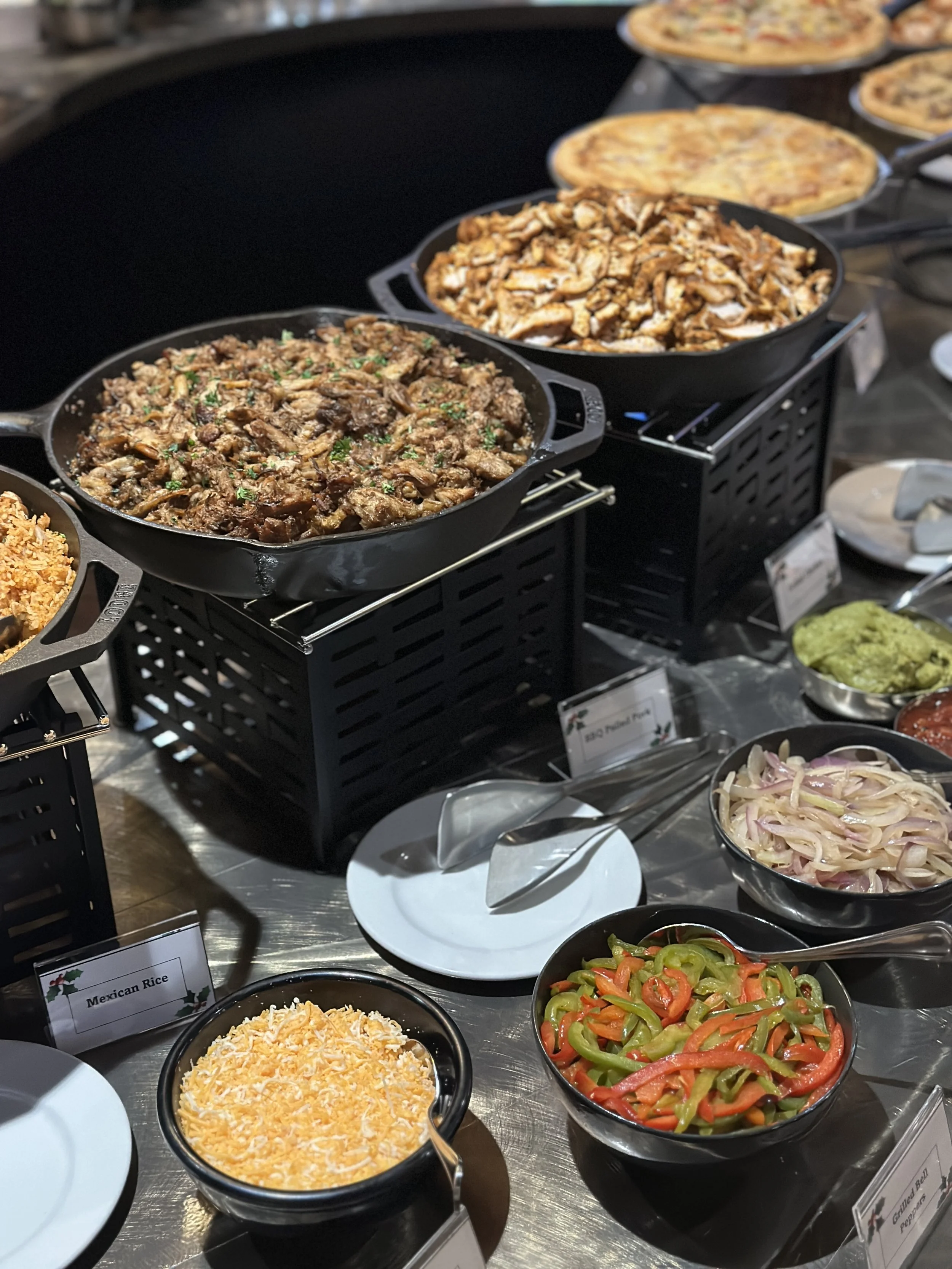 A buffet table with various dishes, including cooked meat, sliced chicken, cheesy rice, and colorful sliced bell peppers and onions, along with slices of pizza in the background.