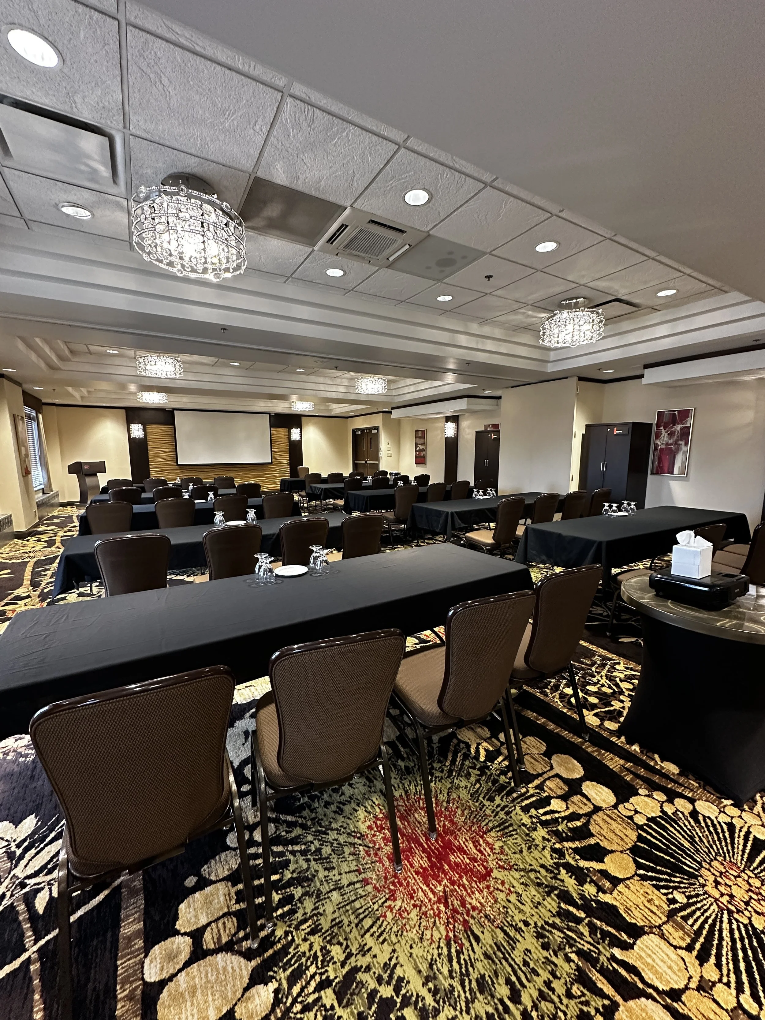 Conference room with black tablecloth-covered tables, brown chairs, and a projector screen at the front, decorated with chandeliers and artistic wall hangings.
