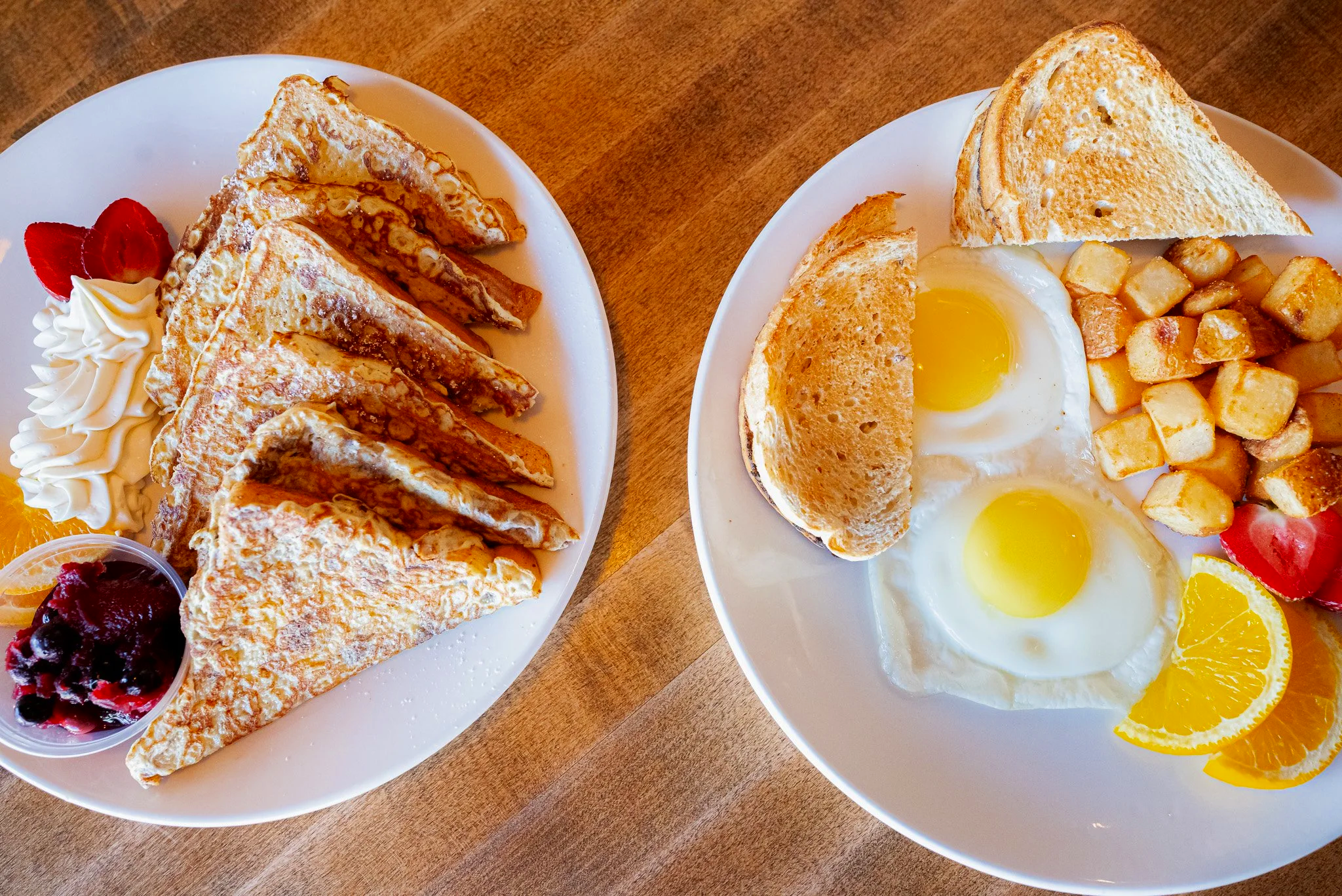 Two white plates of breakfast food on a wooden table: one with caramelized waffle triangles, whipped cream, strawberries, orange slices, and berries; the other with two fried eggs, toasted bread, hash browns, strawberries, and orange slices.