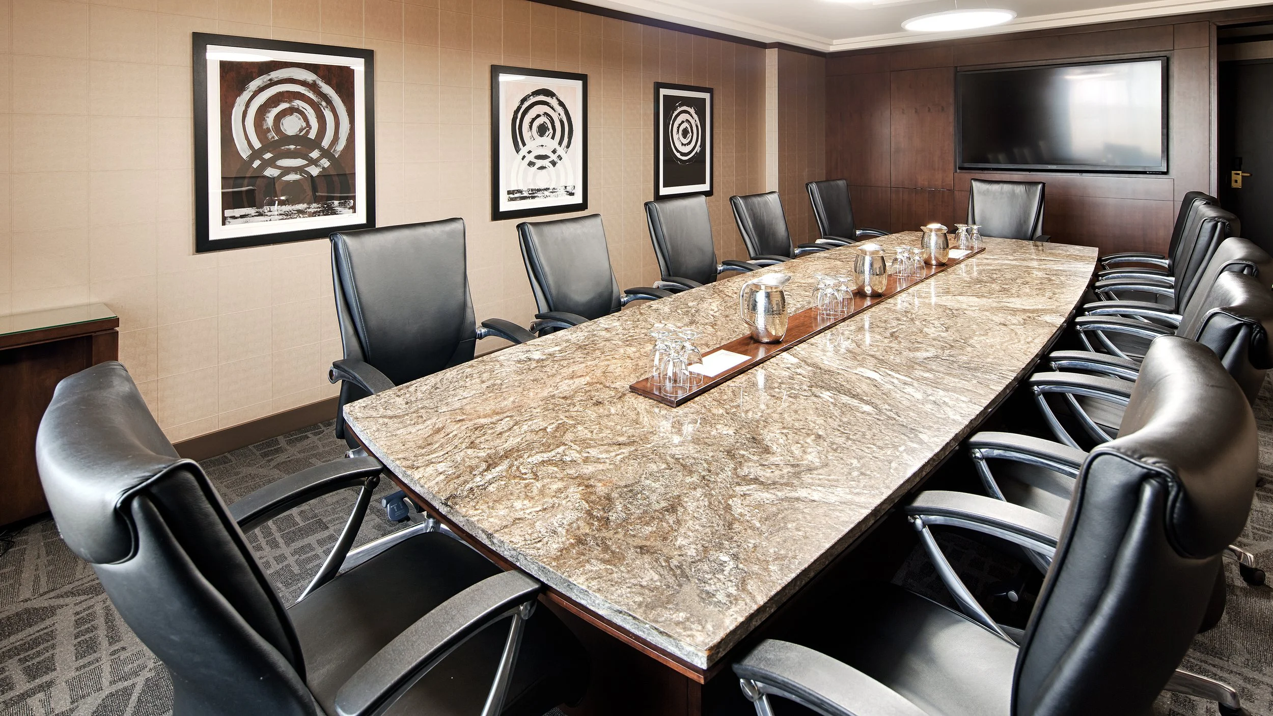 Empty conference room with a long granite table, black leather chairs, abstract art on the wall, a large flat-screen TV, and glasses with water on the table.
