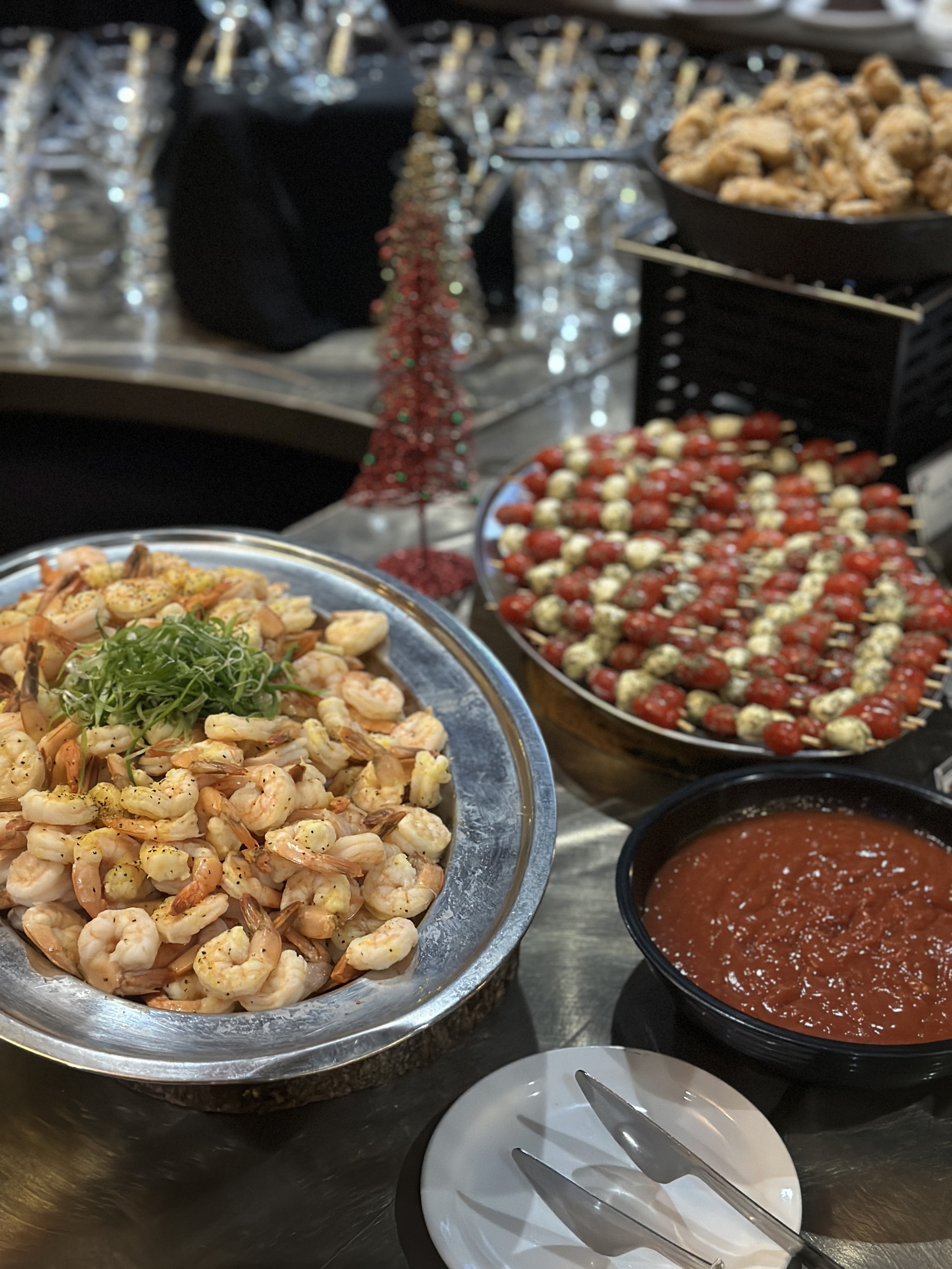 A seafood platter with cooked shrimp topped with green herbs, a bowl of salsa, a tray of cherry tomatoes and burrata skewers, and a bowl of red sauce on a table with a silver tablecloth and holiday decorations.