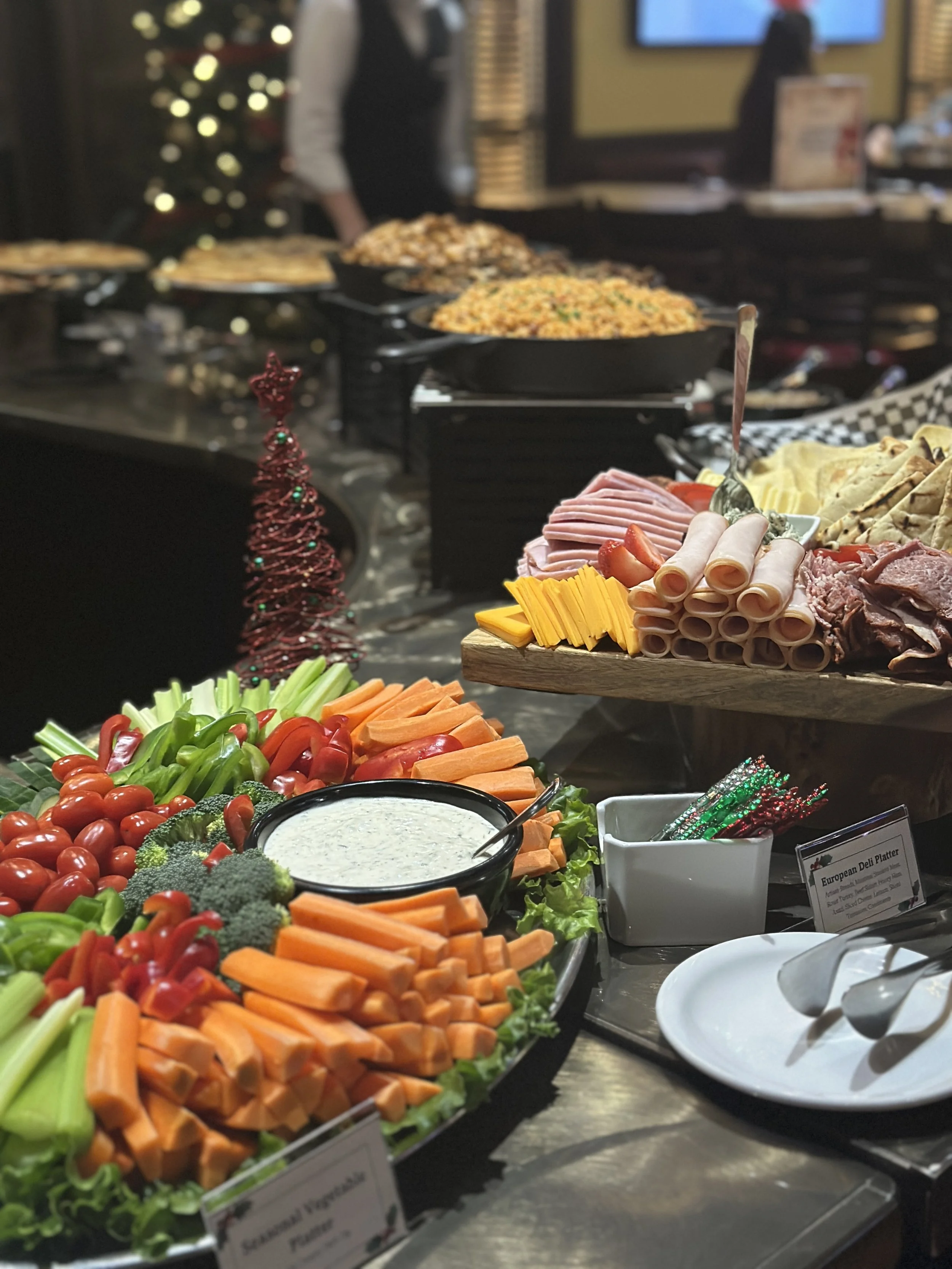 A festive buffet table with vegetables, dips, and assorted cheeses and meats, decorated with small Christmas trees, with more dishes in the background.