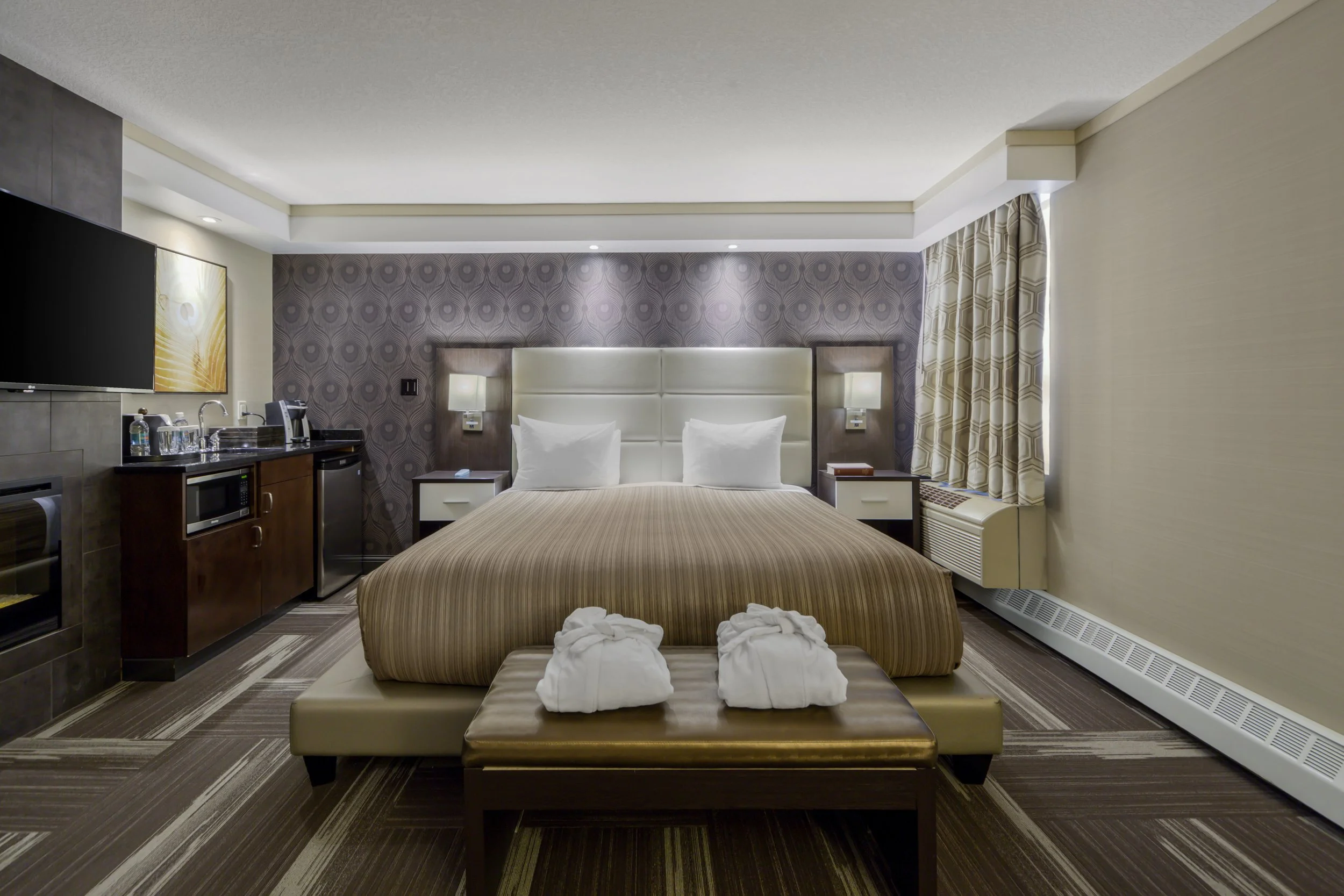 Hotel room with a bed, nightstands, a bench, a microwave, a mini refrigerator, a TV, and a window with curtains.