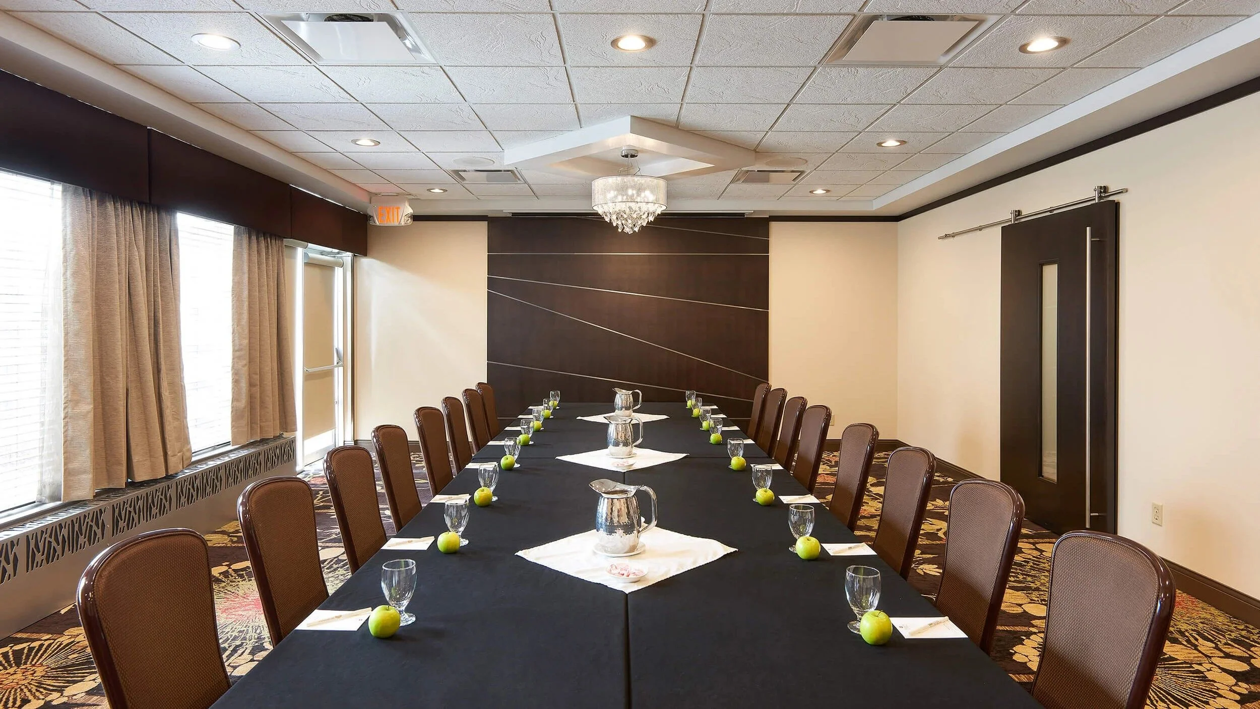 Conference room set up with a long black table, green apples, glasses, pitchers of water, and notepads. There are brown chairs around the table and a chandelier hanging from the ceiling.