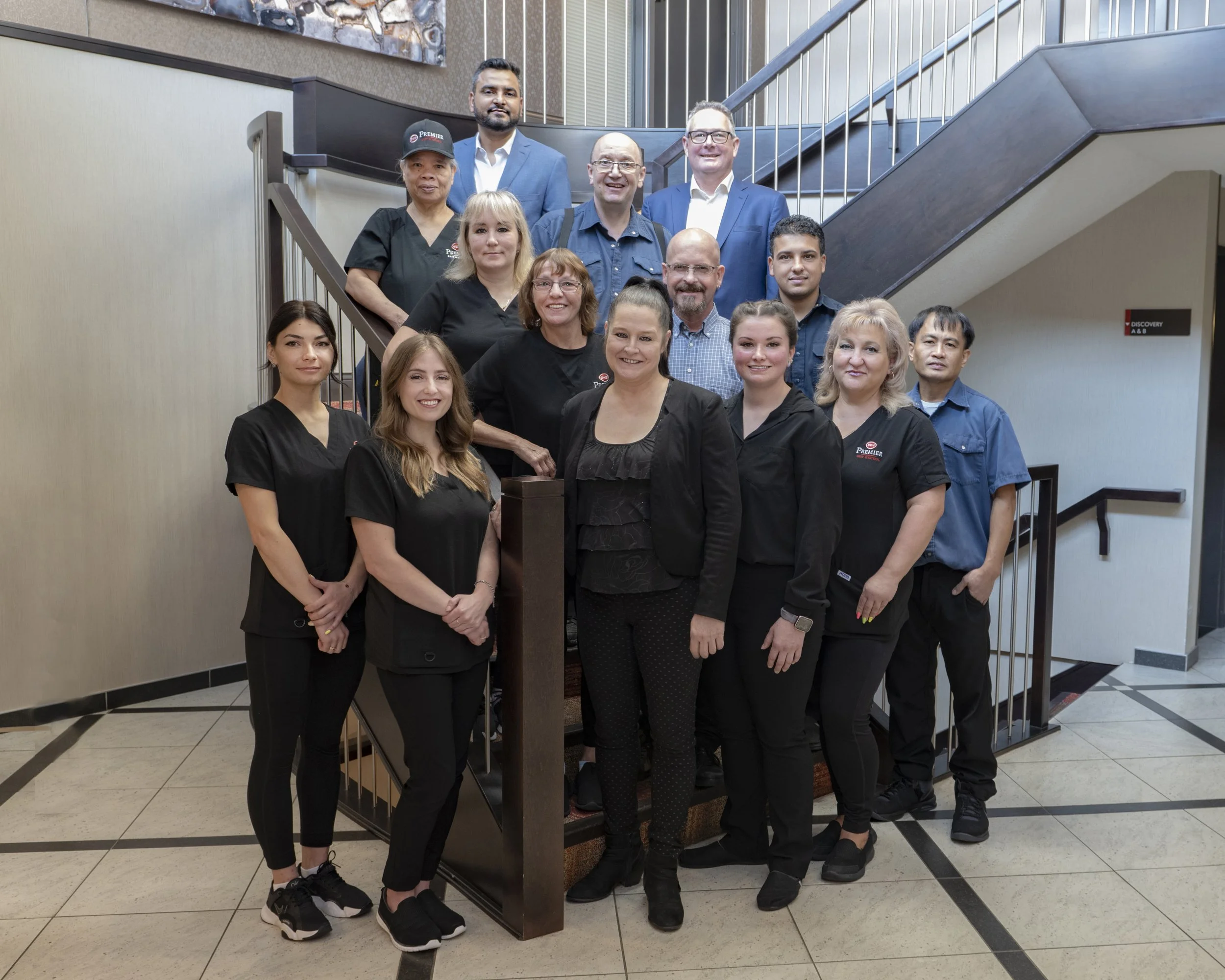 Group of hospitality staff and management posing on a staircase in a hotel or restaurant. The group includes both men and women in professional attire, with some wearing black scrubs and others in blue blouses or suits.