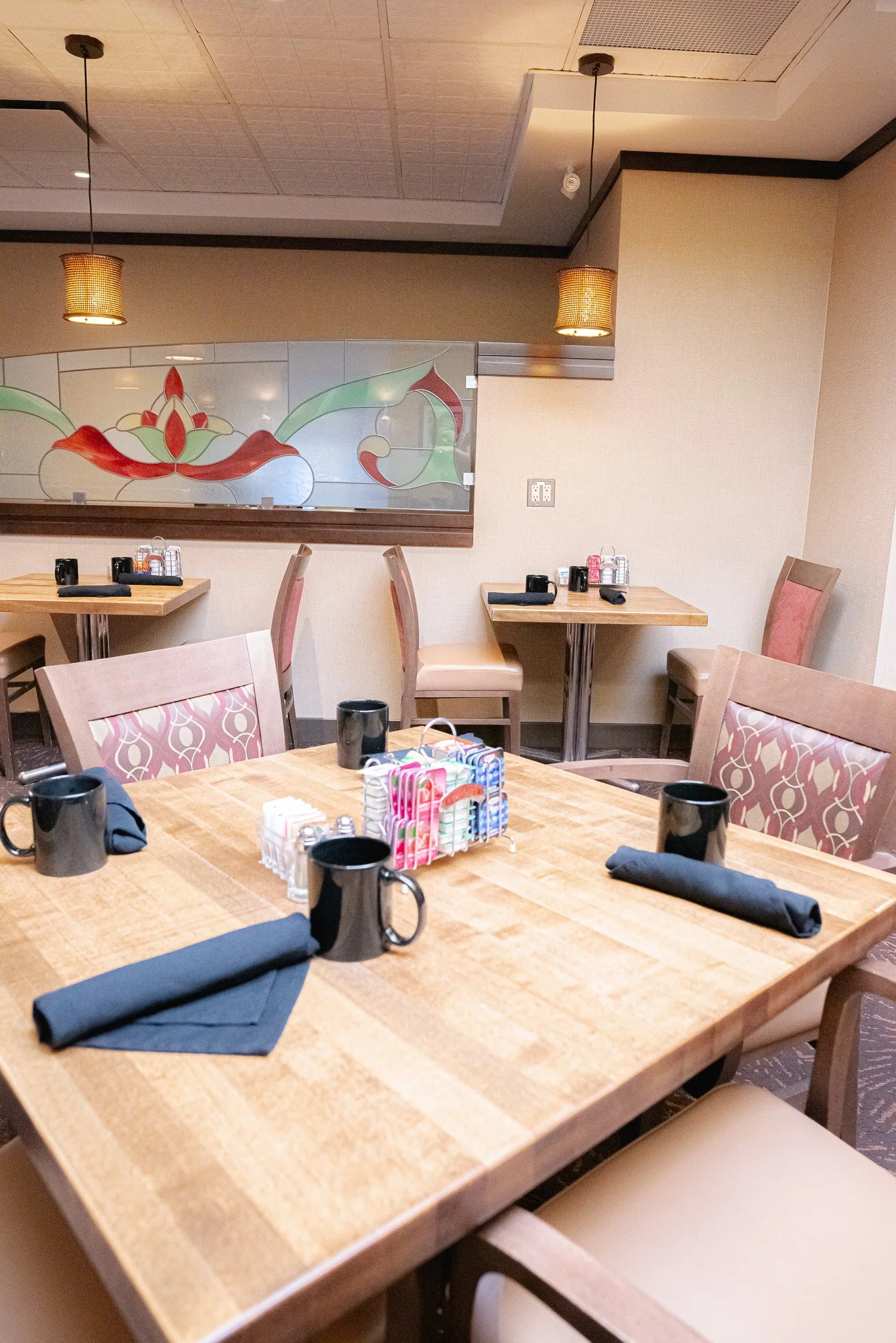 A restaurant dining area with a wooden table set with black mugs, black napkins, a condiment caddy, and a small decorative item. There are multiple chairs around the table, some with patterned upholstery. In the background, there are two smaller tables with black mugs, napkins, condiments, and chairs. The wall features a decorative glass panel with a floral design, and the ceiling has hanging lights and a vent.