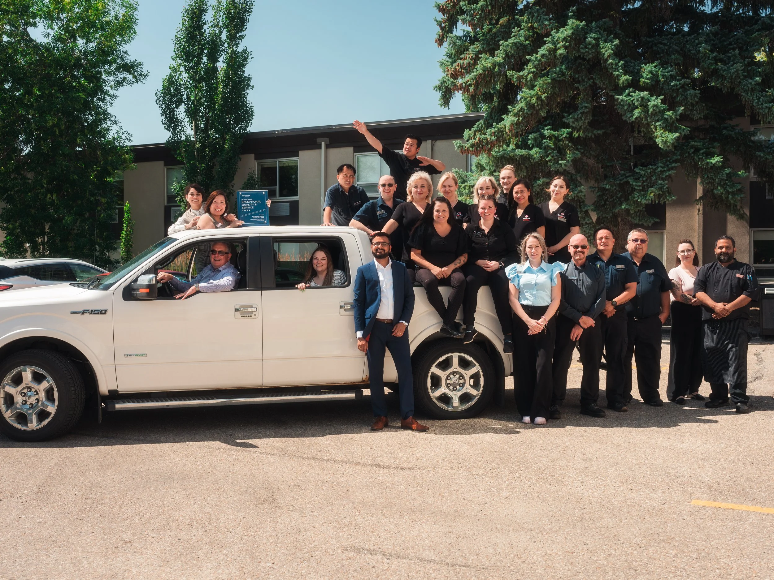 Group of people gathered around and on a white pickup truck, posing for a photo outdoors in front of a building and trees, some smiling, some sitting on the truck's hood and roof, others standing nearby