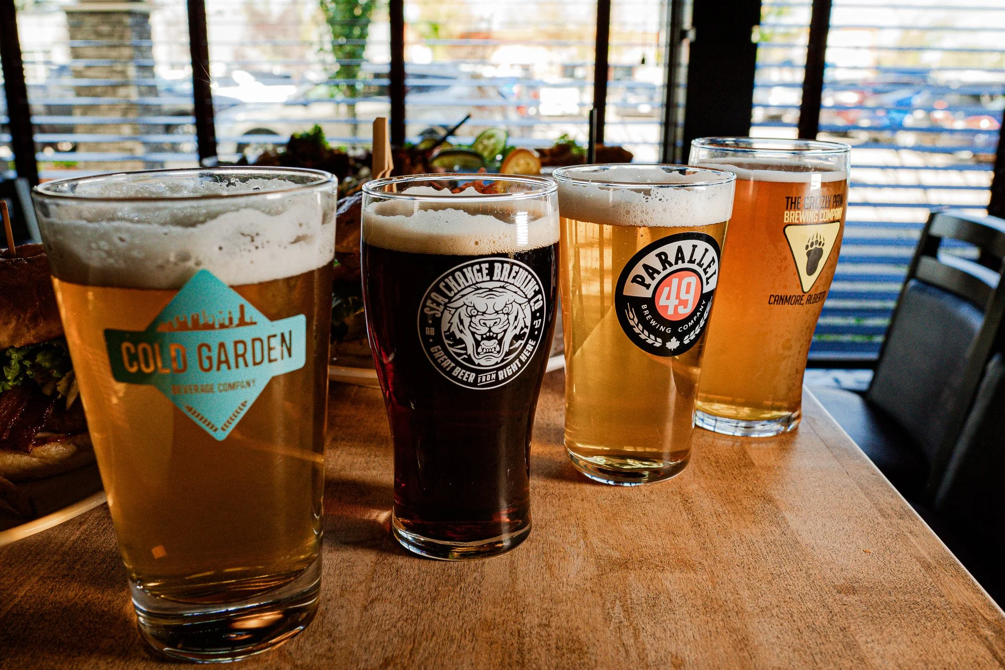 Four glasses of craft beer on a wooden table in a restaurant, with a food platter in the background.