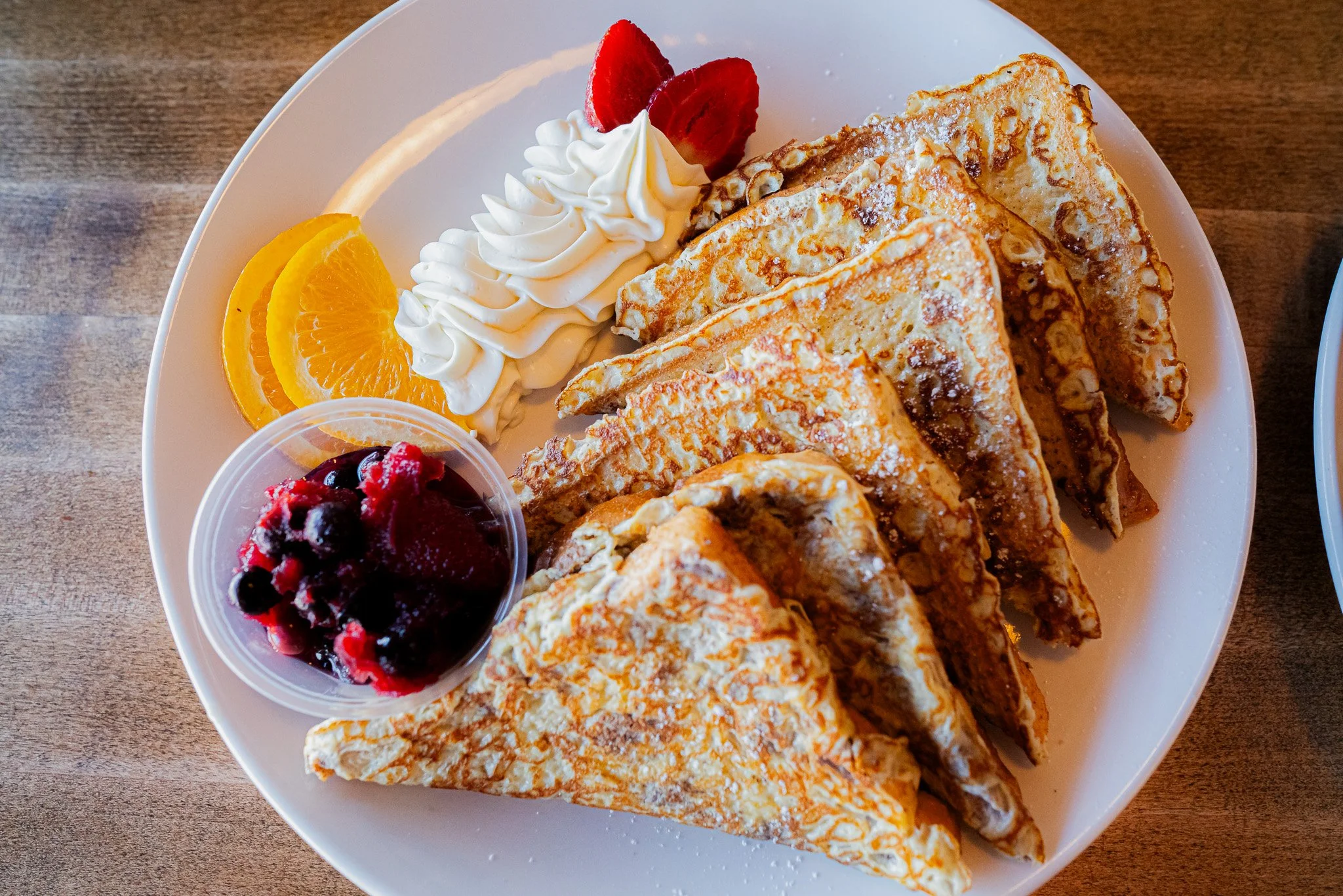 A white plate with five folded french toast slices dusted with powdered sugar, garnished with whipped cream, strawberry and orange slices, and a side of mixed berry compote.