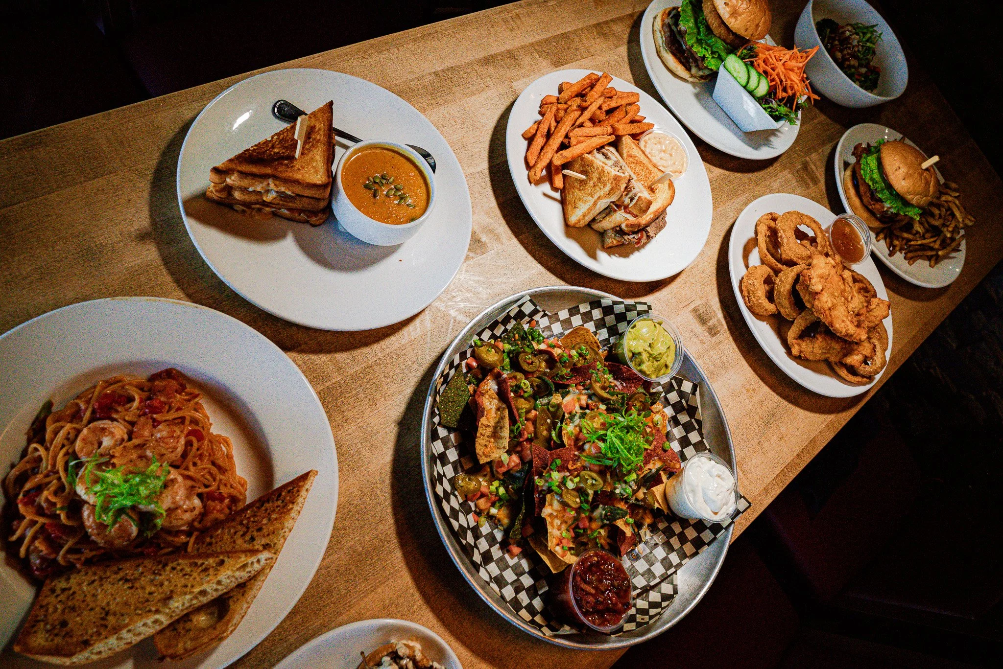 Assorted plates of food including sandwiches, salads, fried onion rings, fries, pasta, and soups on a wooden table.