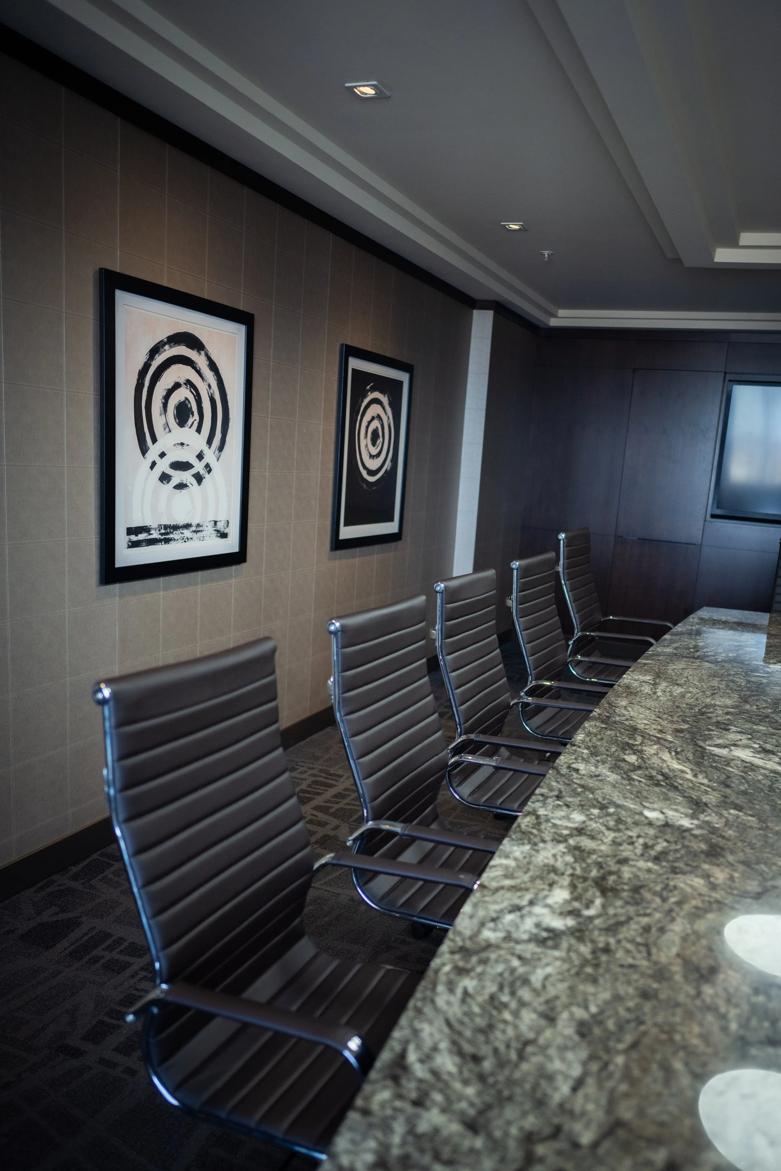 Empty conference room with five black leather chairs arranged along a granite table, and framed abstract black and white artwork on the beige tiled wall.