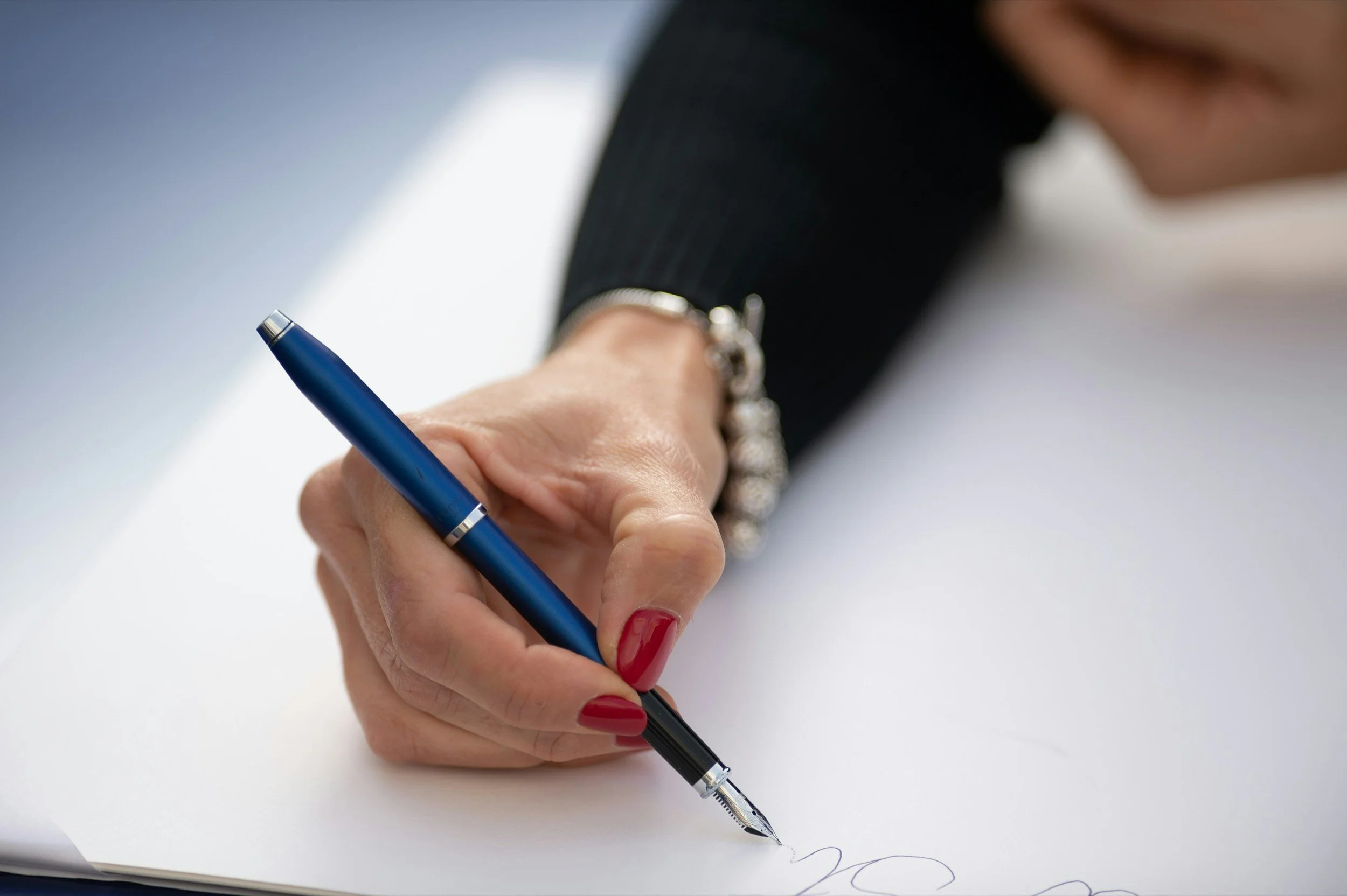 Close-up of a woman's hand with red painted nails, holding a blue fountain pen and writing on a white piece of paper.