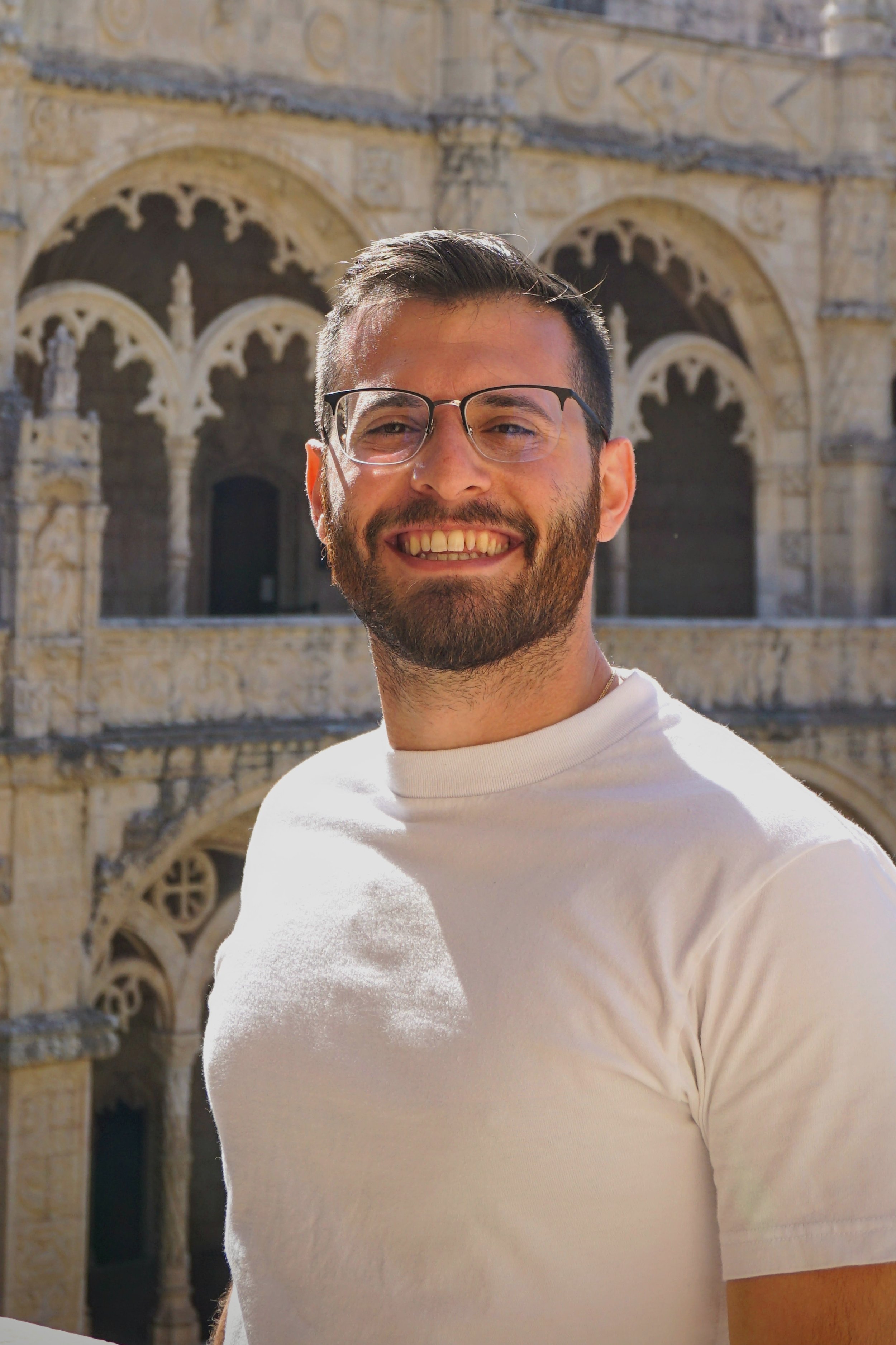 A smiling man with glasses and a beard standing outdoors in front of a historic, stone building with arched windows and intricate architectural details.