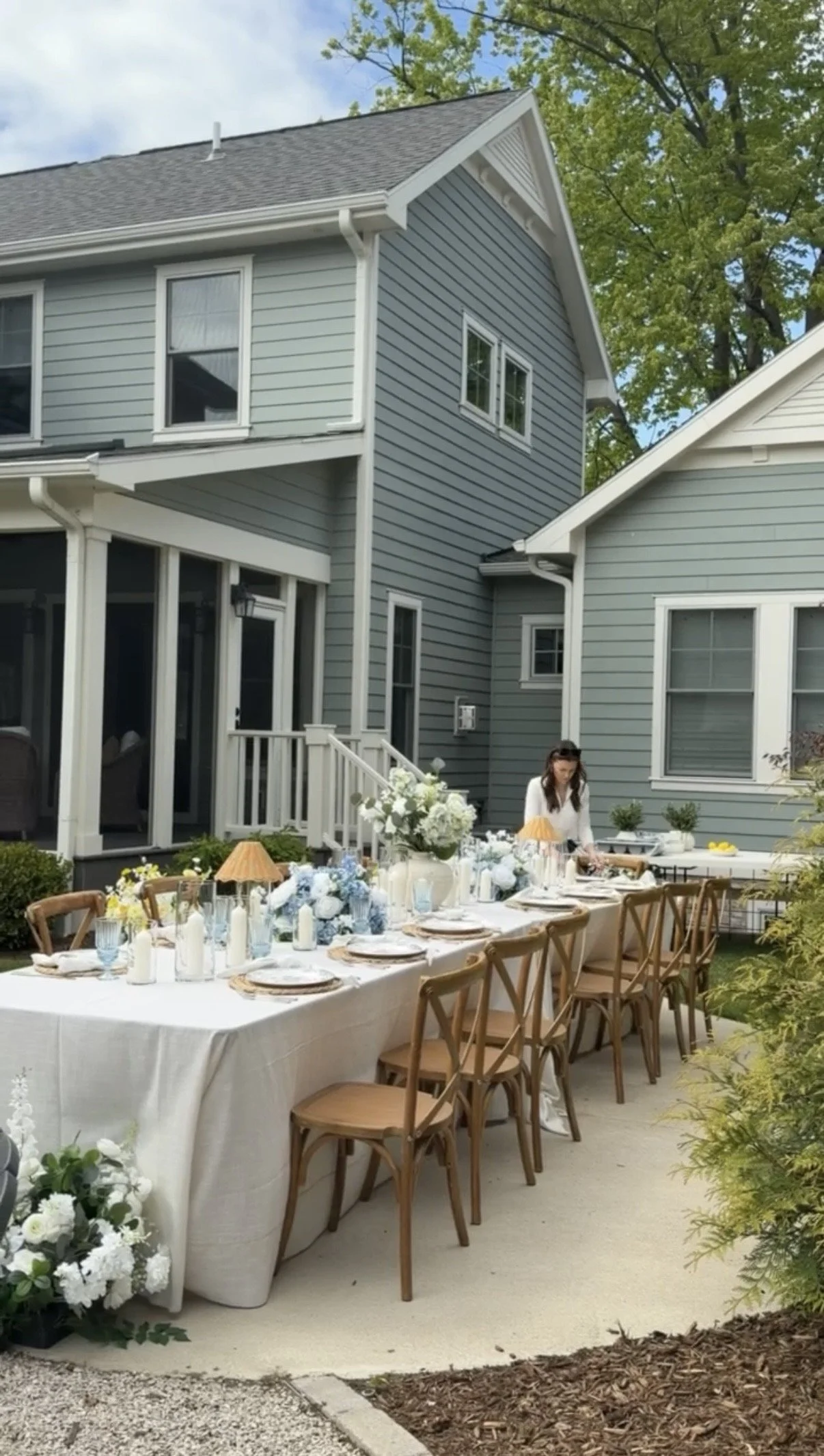 A backyard with a large rectangular dining table set for a meal, decorated with white and blue flowers and candles. A woman is standing at the table, possibly preparing food or setting up. The house in the background is blue with white trim, multiple