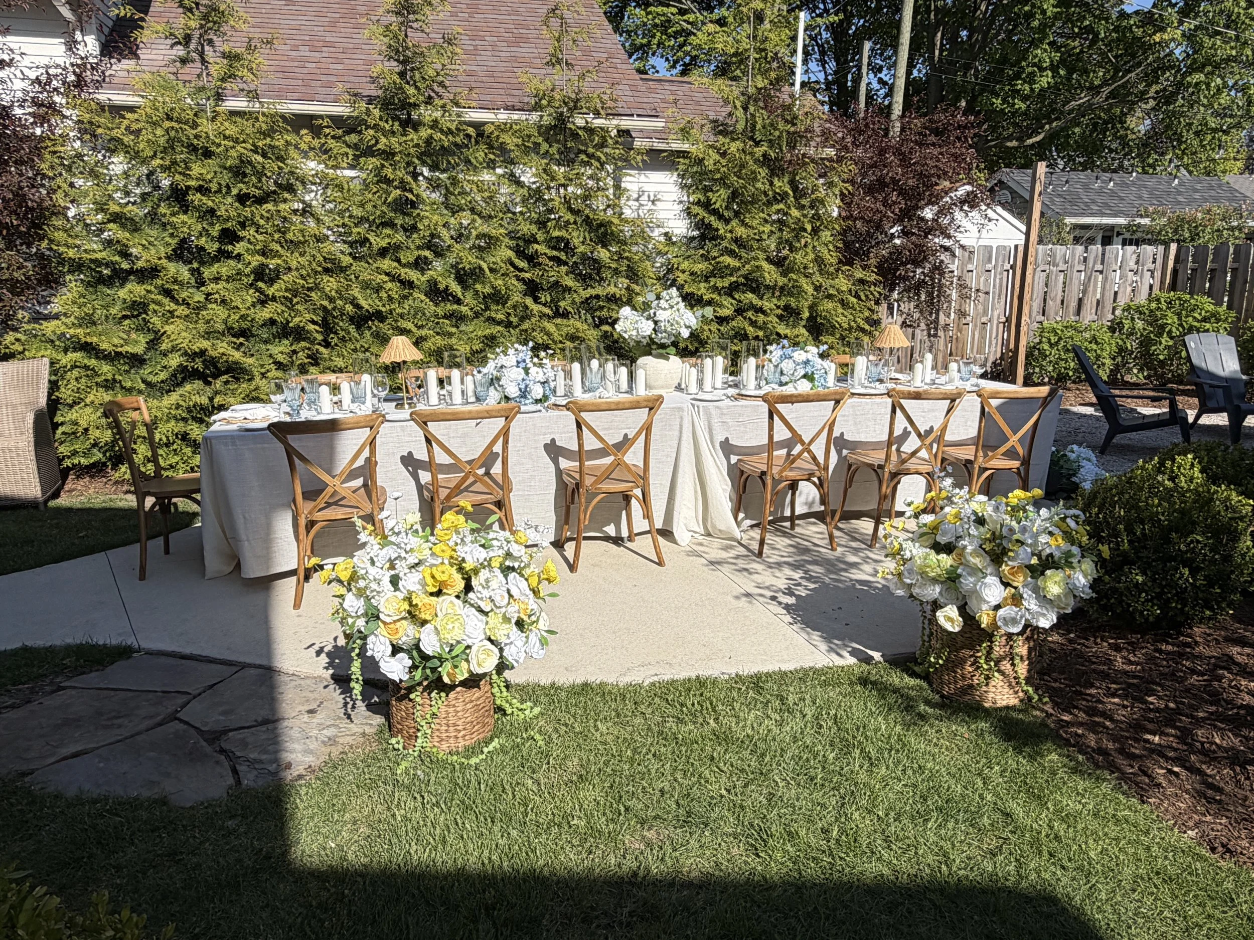 An outdoor dining table decorated with white and blue flowers, candles, and small lamps, set up on a patio in a garden with green trees and a wooden fence in the background.