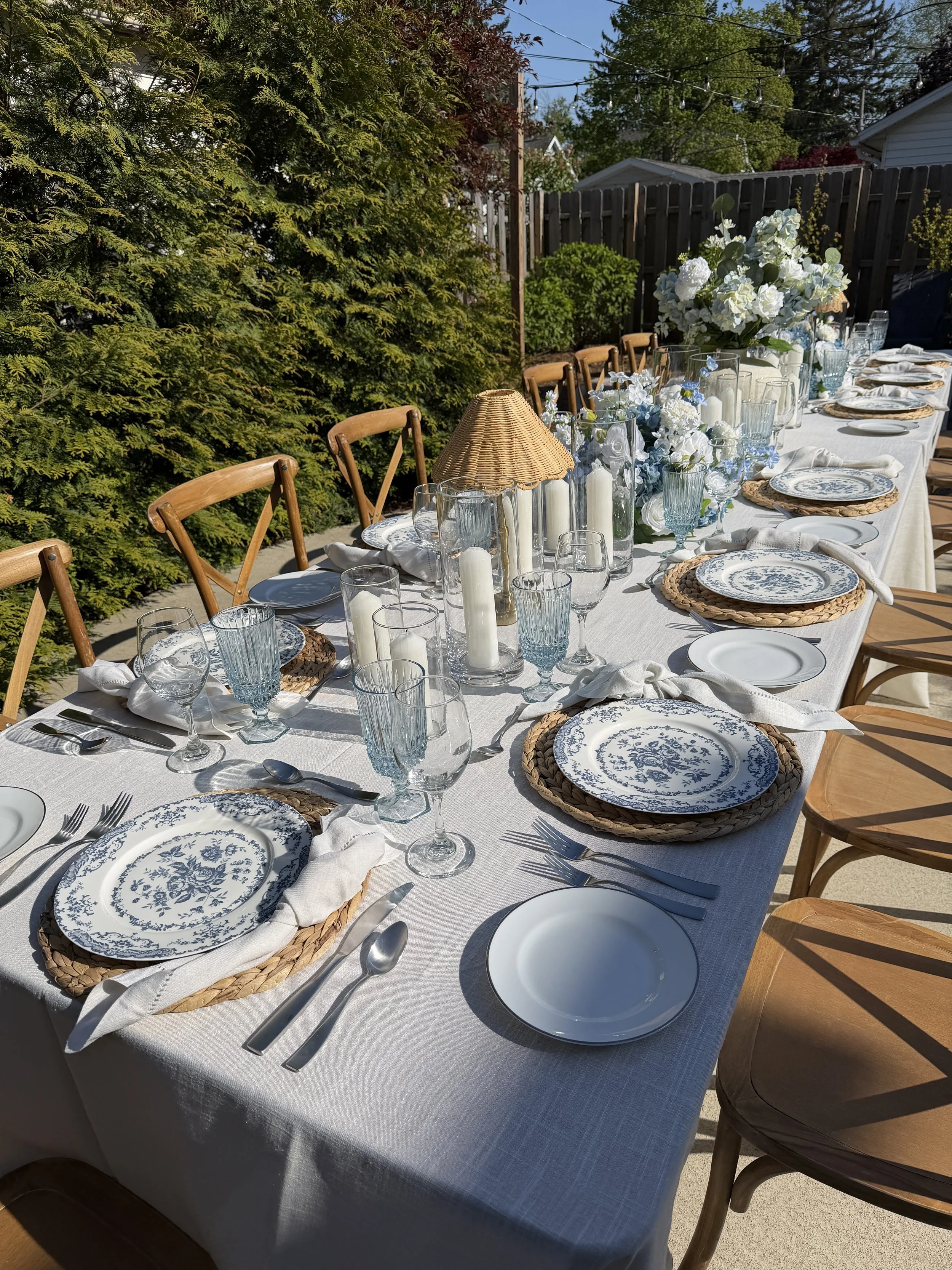 A long outdoor dining table set for a meal, with blue and white dishware, glasses, candles, and a floral centerpiece on a white tablecloth. Wooden chairs surround the table, which is placed in a backyard with green hedges and trees under a clear sky.