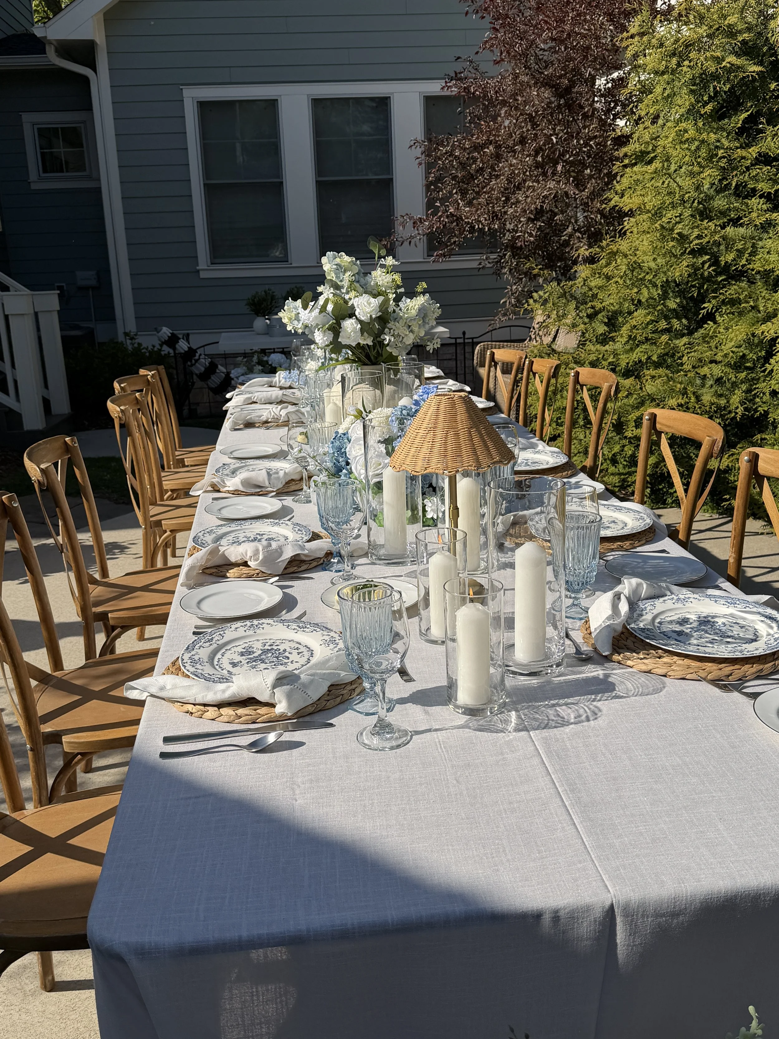 Outdoor dining table set for a meal with white tablecloth, floral centerpiece, candles, glasses, plates, and utensils, surrounded by wooden chairs, in a patio area with trees and a house in the background.