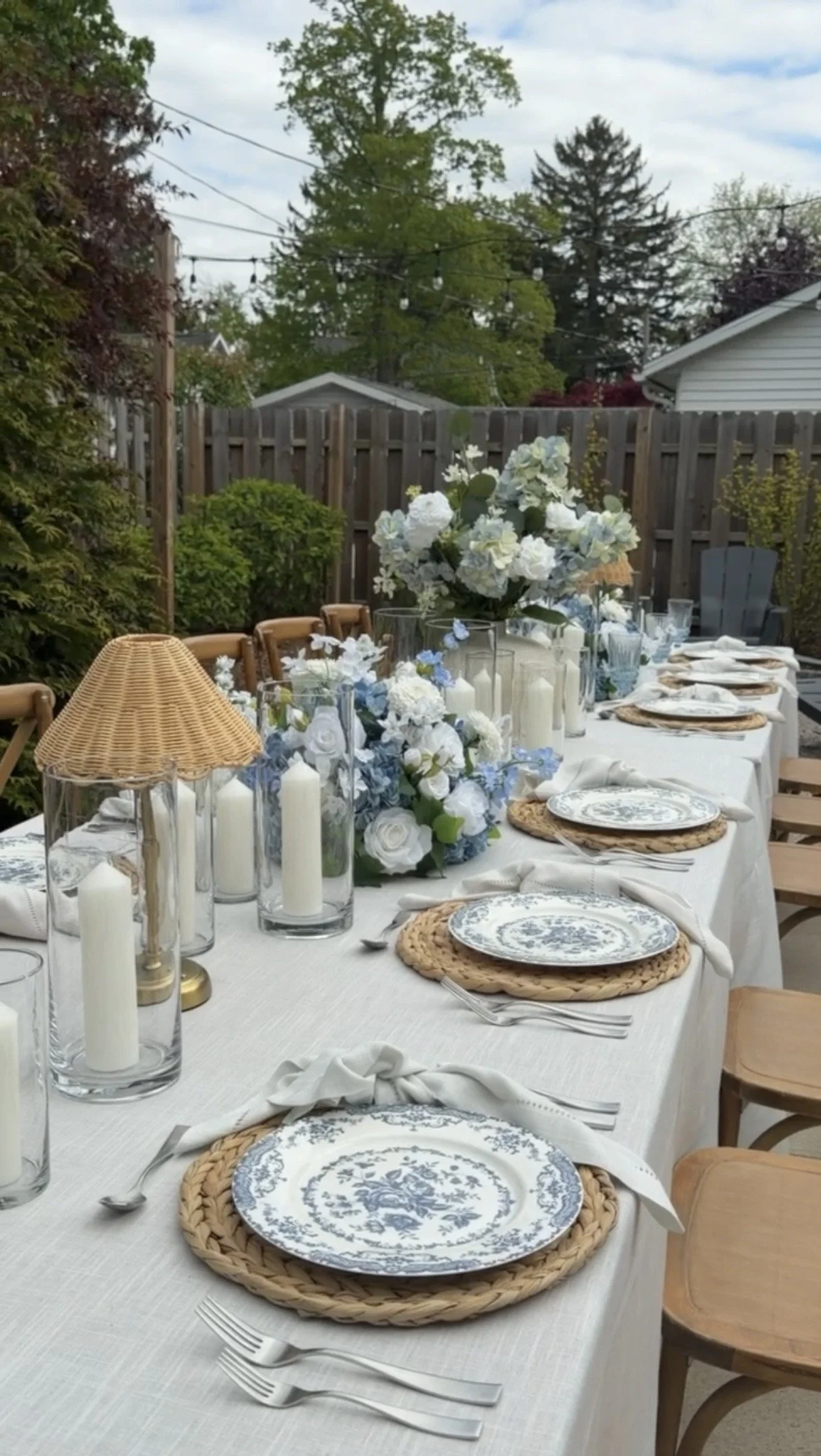 Outdoor dining table decorated with blue and white floral arrangements and candles, set with plates, napkins, and utensils for a gathering.