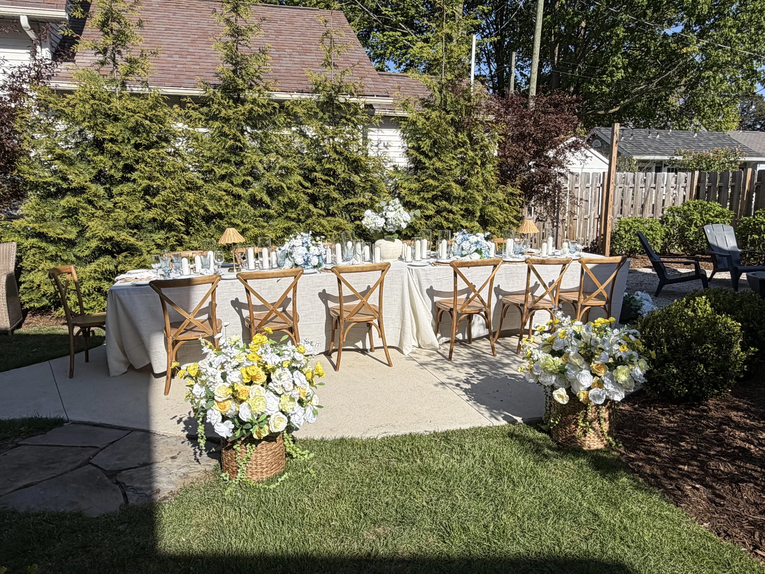 An outdoor celebration setup with a long table covered in a beige tablecloth, decorated with white floral centerpieces and candles. The table is surrounded by wooden chairs. In the foreground, there are large woven baskets filled with white and yello
