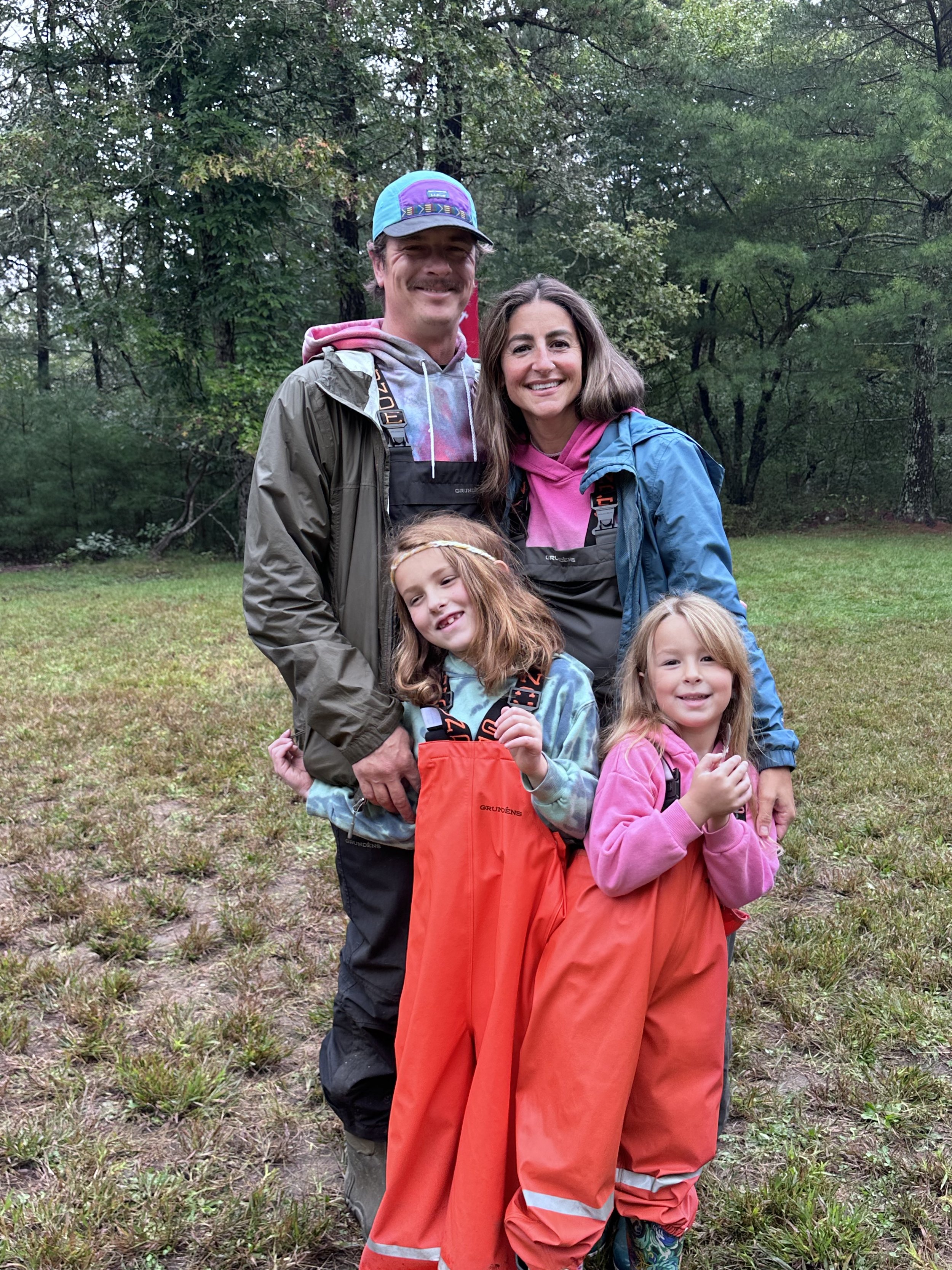 A happy family of four, including two adults and two young girls, standing outdoors in a grassy, wooded area, dressed in rain gear.