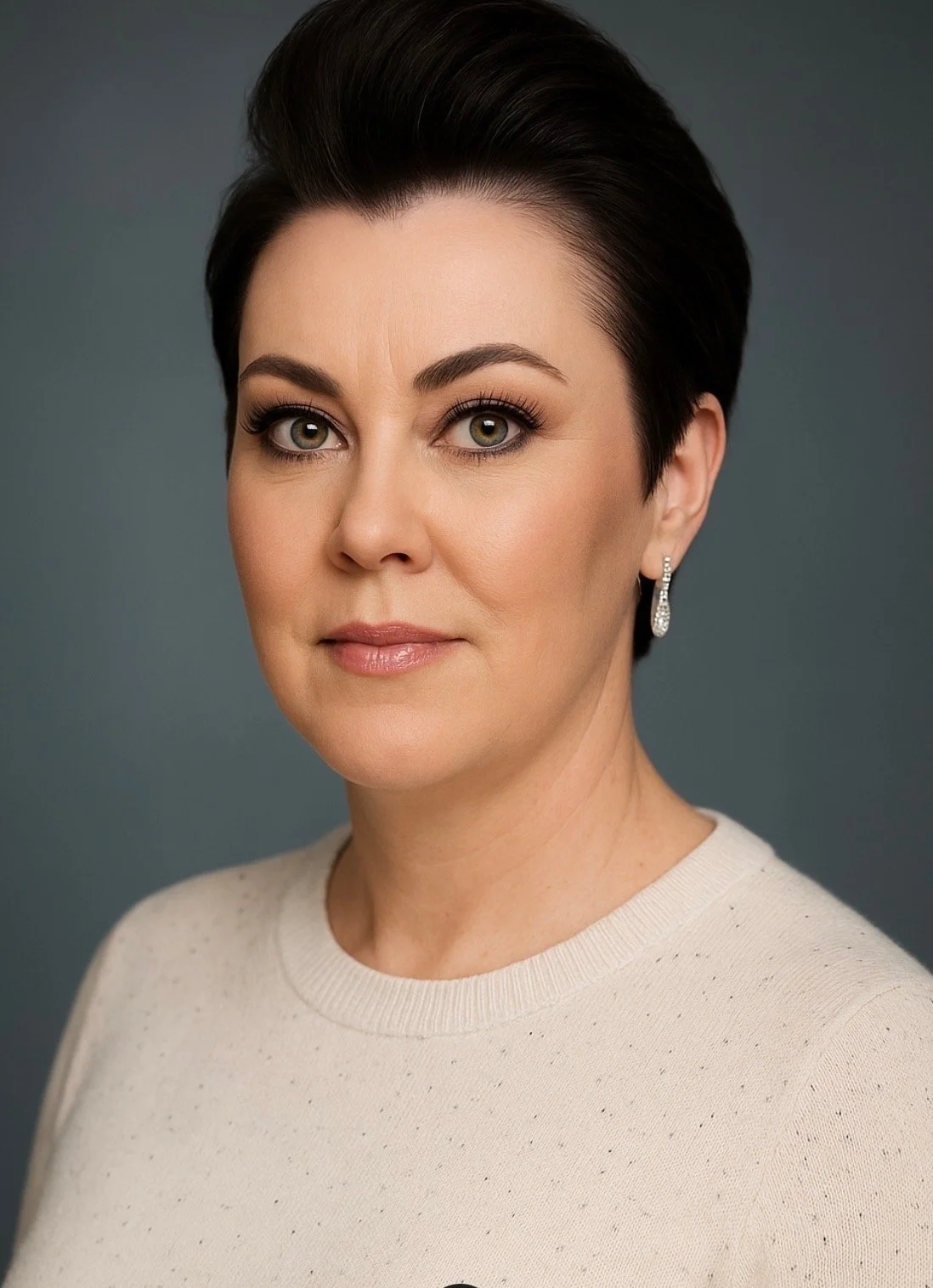 A woman with short dark hair, wearing pearl earrings and a beige sweater, looking directly at the camera against a plain background.