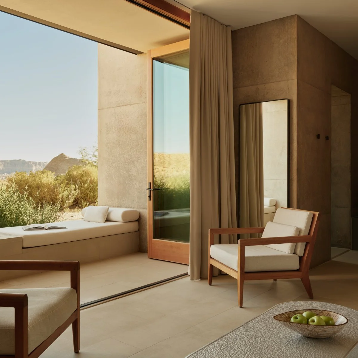 Modern living room with large glass door opening to desert landscape, featuring minimalist furniture and neutral color palette.