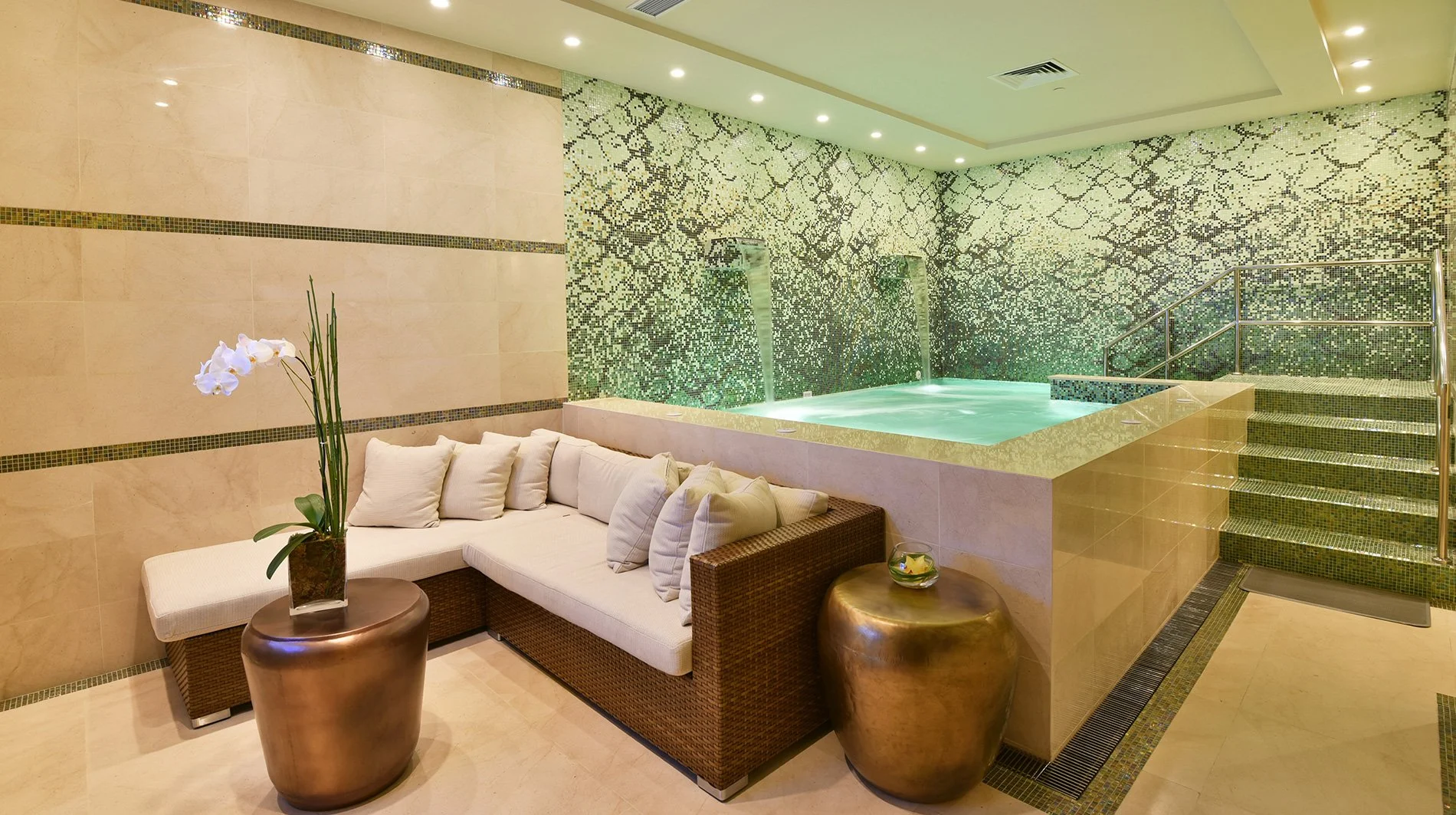 Indoor spa area with a jacuzzi, mosaic wall, beige tiled walls, a white cushioned L-shaped sofa with multiple pillows, a large brass vase with a white orchid on a small round brass table, and warm ambient lighting.