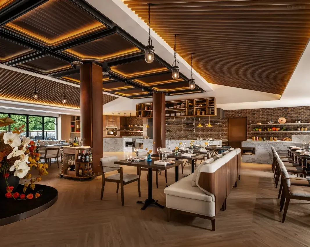 Inside a modern restaurant with wooden ceiling, large windows, and elegant decor. There are tables set with flowers, glasses, and napkins, along with cushioned seating and a brick wall in the background.