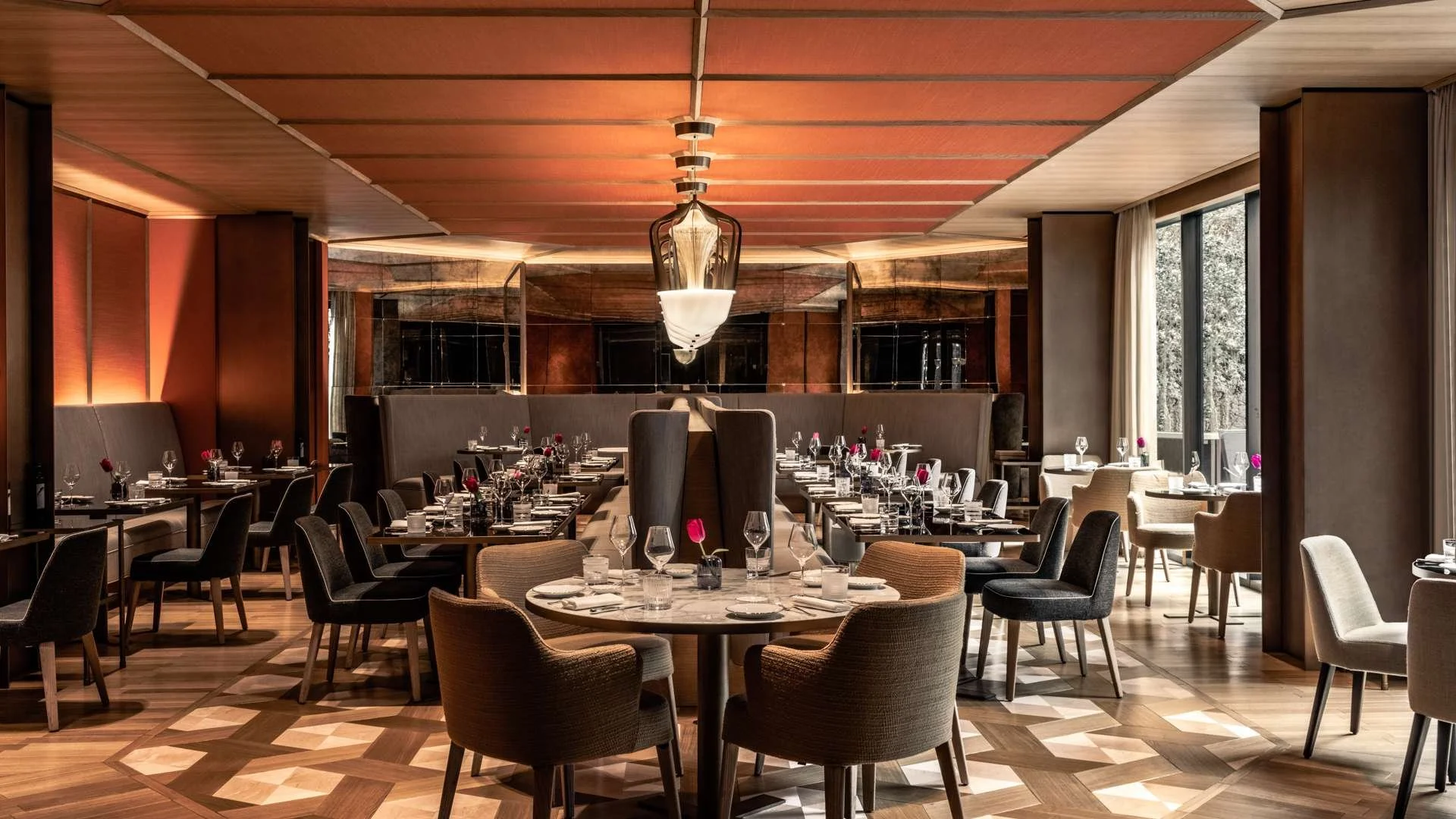 Elegant restaurant dining area with round and rectangular tables set with wine glasses, silverware, and pink roses. Modern chandelier hanging from a wooden ceiling, large windows letting in natural light, and a geometric-patterned wooden floor.