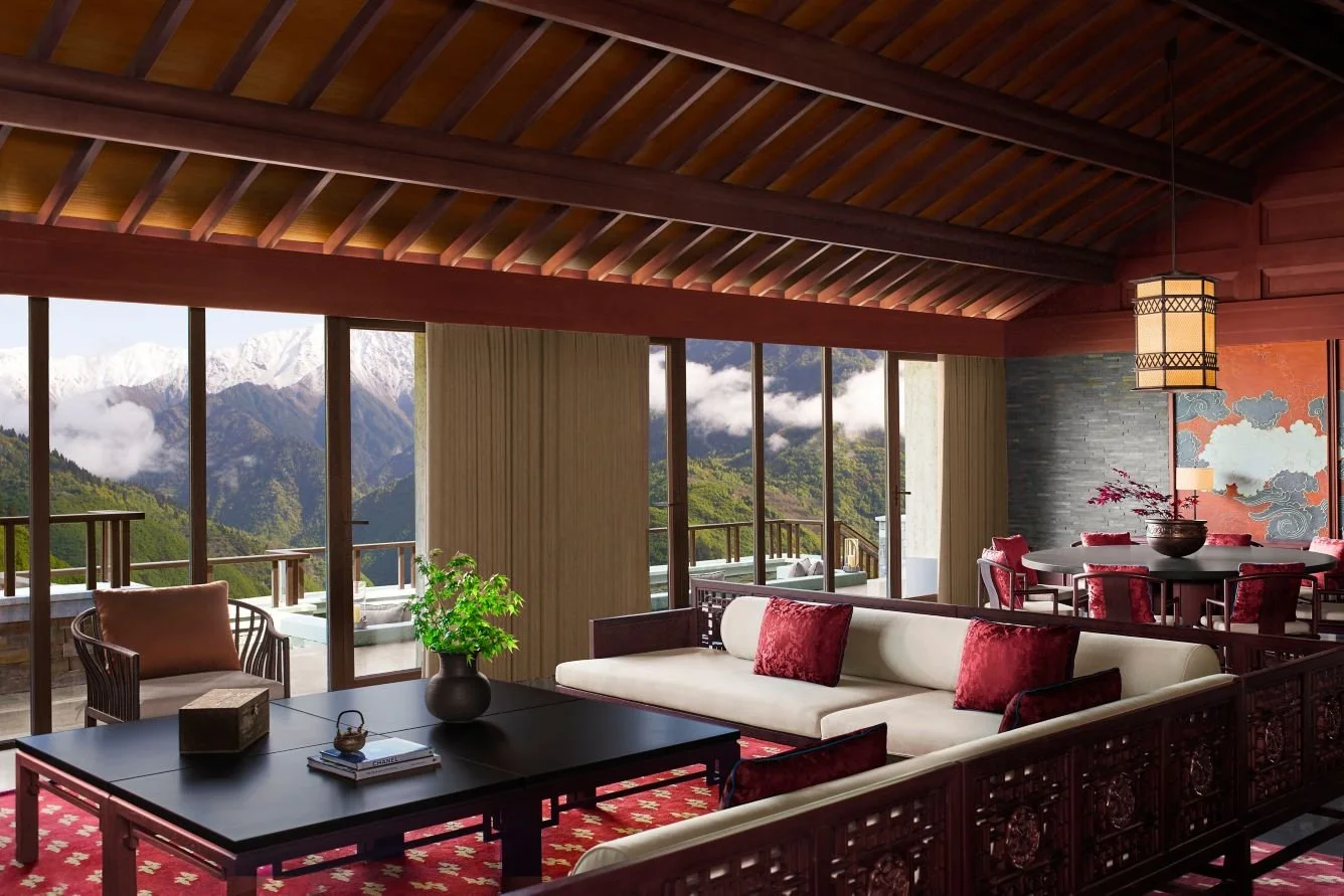 Living room with large glass doors opening to mountain and forest view, decorated with Asian-inspired furnishings, including a white sofa with red cushions, a black coffee table with a potted plant, and a dining area with red upholstered chairs and a
