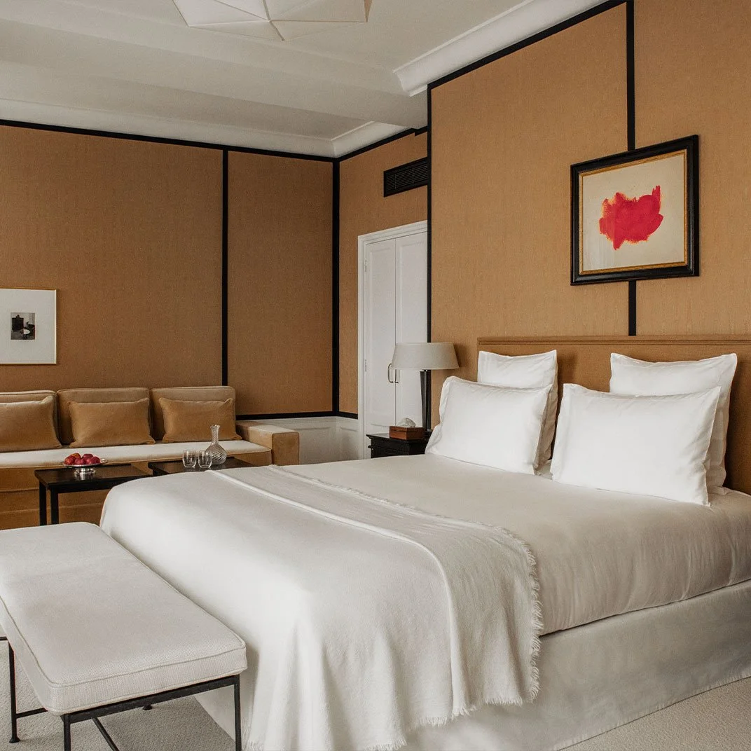 A hotel room with a large bed, white linens, and four pillows. There is a beige sofa with pillows and a small black table with glassware and apples. Artwork hangs above the bed and the room has beige and brown decor.
