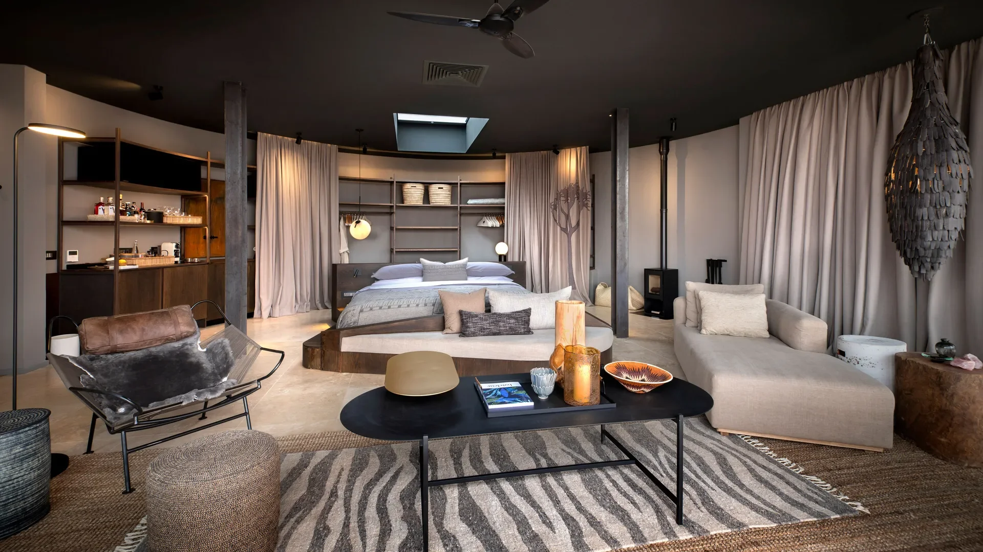 A spacious modern bedroom with a large bed, sitting area, and decor in neutral tones, featuring curtains, a black ceiling, and open shelving.