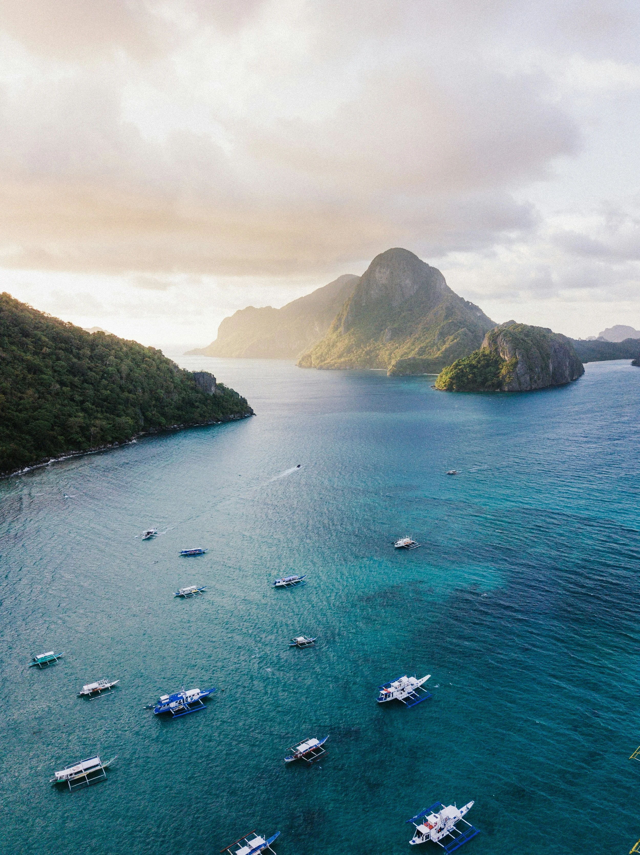 Aerial view of a bay with multiple boats floating on blue water, surrounded by lush green hills and mountains, under a partly cloudy sky.