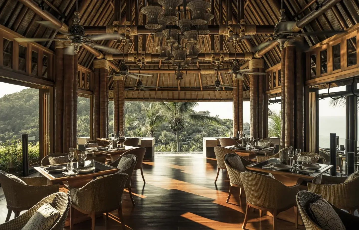 A tropical restaurant with outdoor view of lush greenery and palm trees through large windows, featuring wooden tables and wicker chairs, and a high bamboo ceiling.