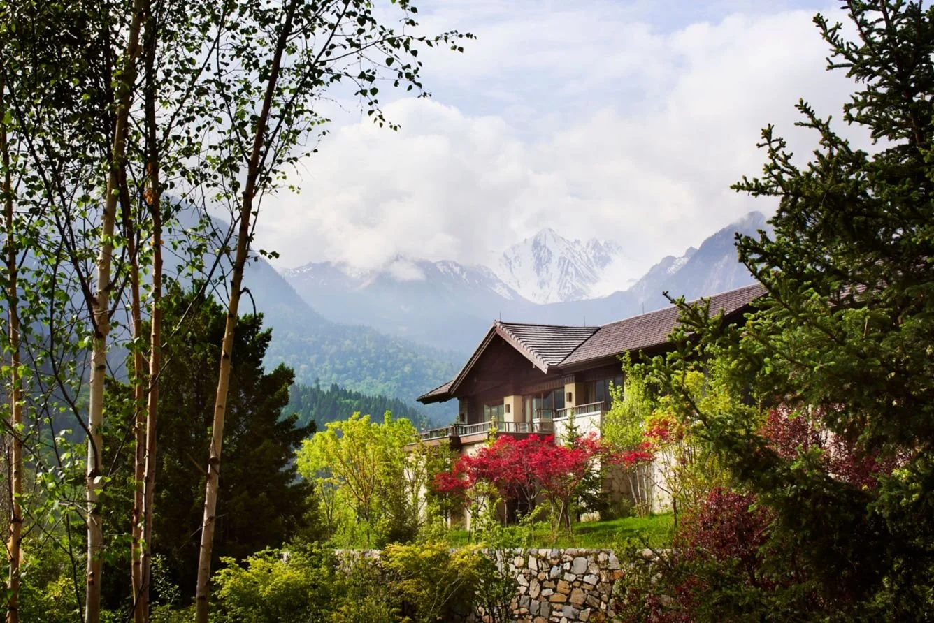 A house surrounded by colorful trees and lush greenery with snow-capped mountains in the background.
