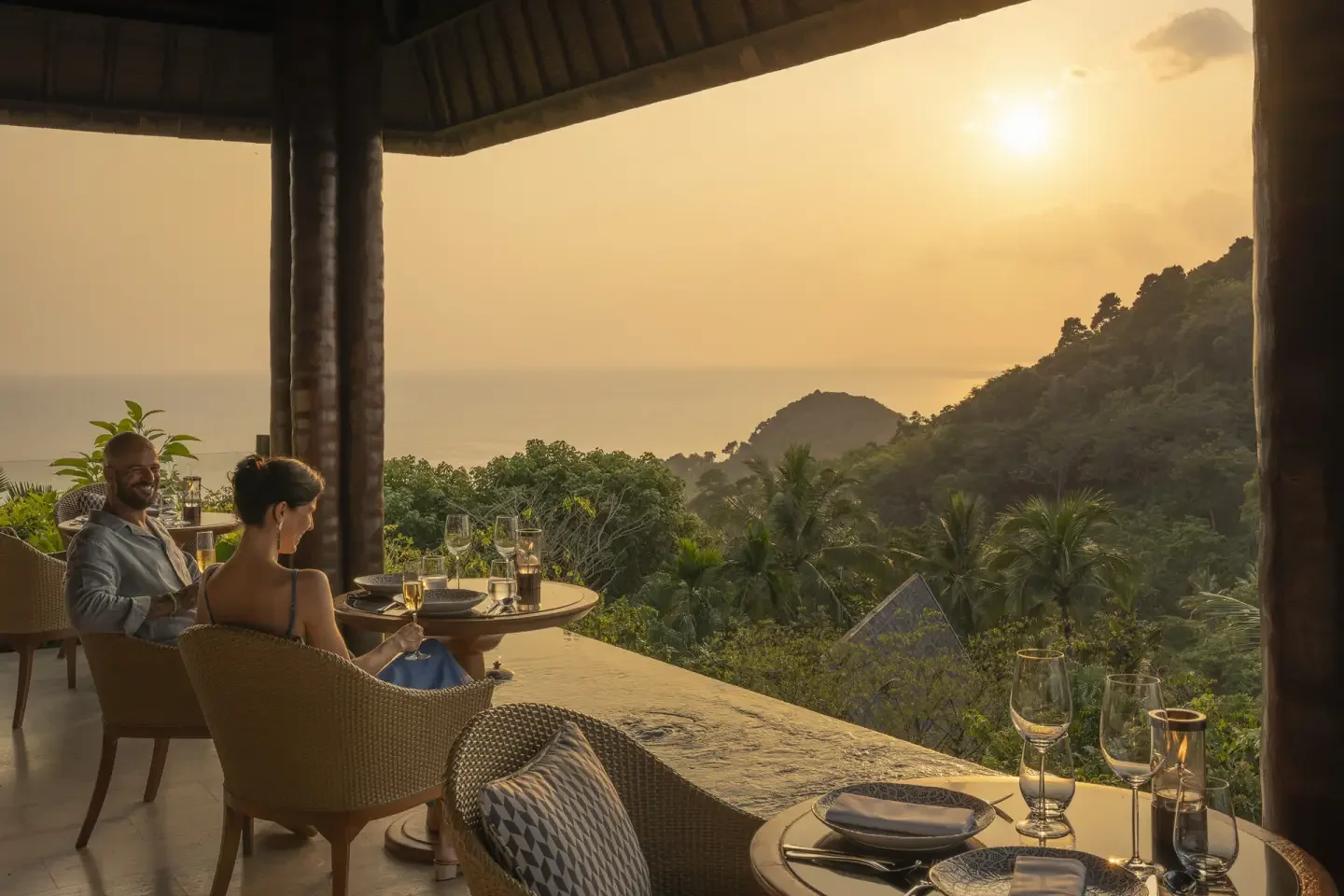 Two people sitting at a table on a balcony overlooking a lush tropical landscape and the ocean during sunset, with empty wine glasses and table settings.