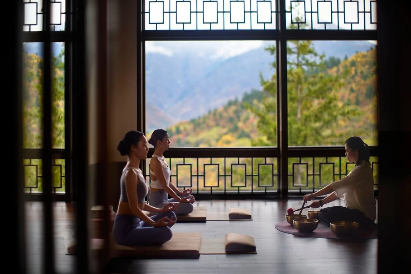 Three women sitting cross-legged on yoga mats in a meditation class, guided by an instructor, with large windows revealing a scenic mountain landscape outside.