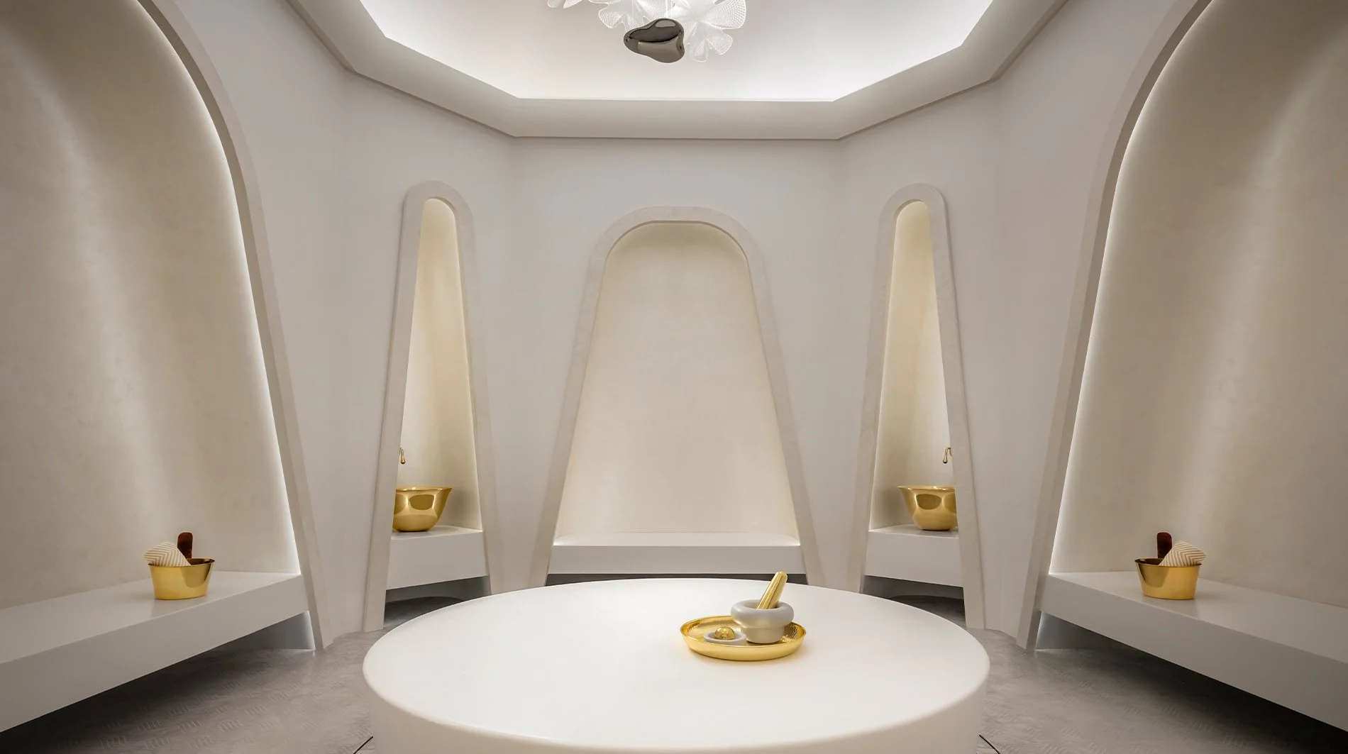 Minimalist white room with built-in shelves, a round table in the center with a mortar and pestle, and decorative items in gold and white.