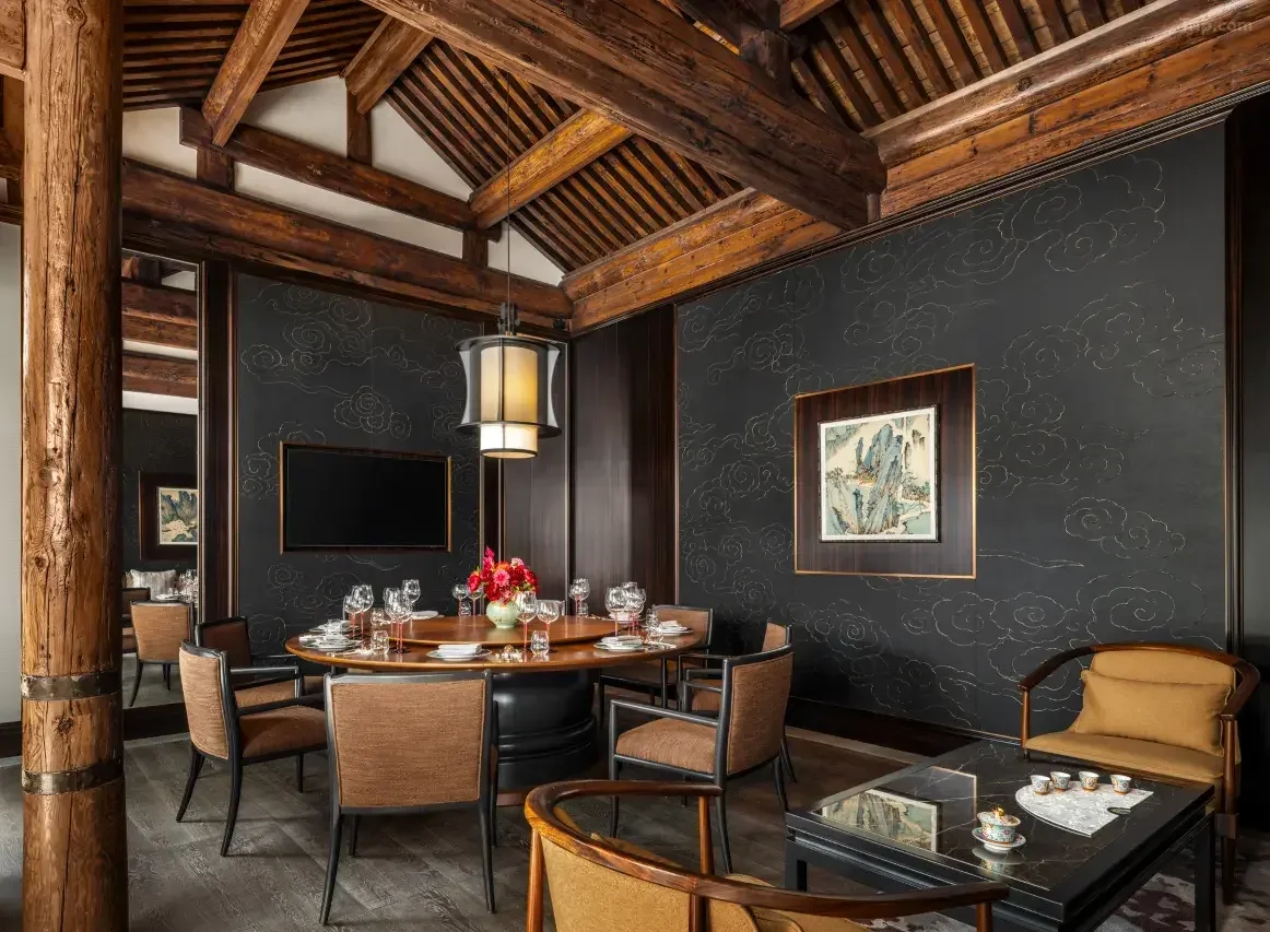 Elegant dining room with dark wood paneling, a round wooden table set with glassware and a floral centerpiece, surrounded by cushioned chairs, a black accent wall with chalk art, and a yellow cushioned armchair and a black coffee table with tea cups.