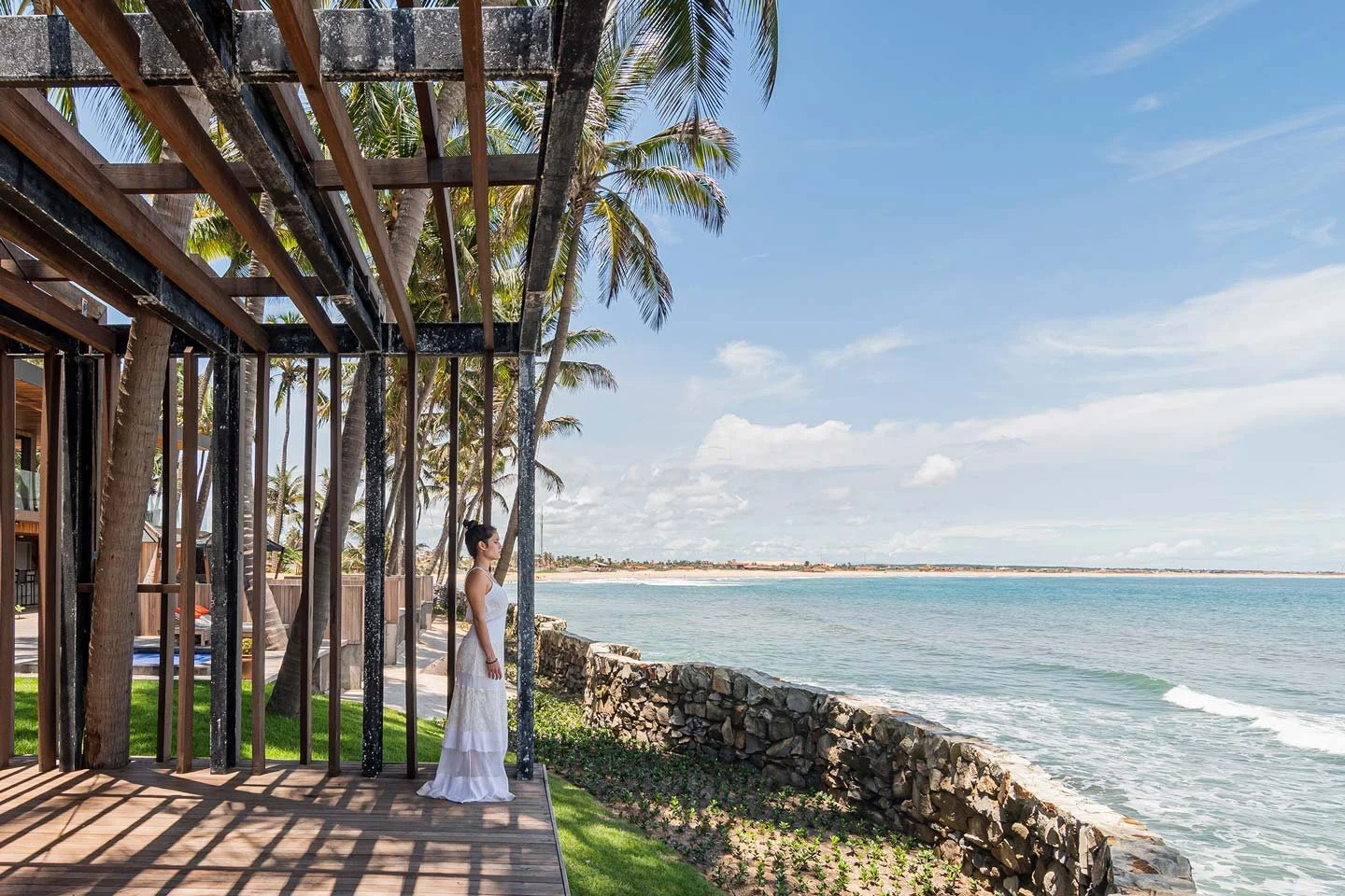 A woman in a long white dress standing on a wooden deck near the ocean, with palm trees and thatched cottages in the background, under a partly cloudy sky.