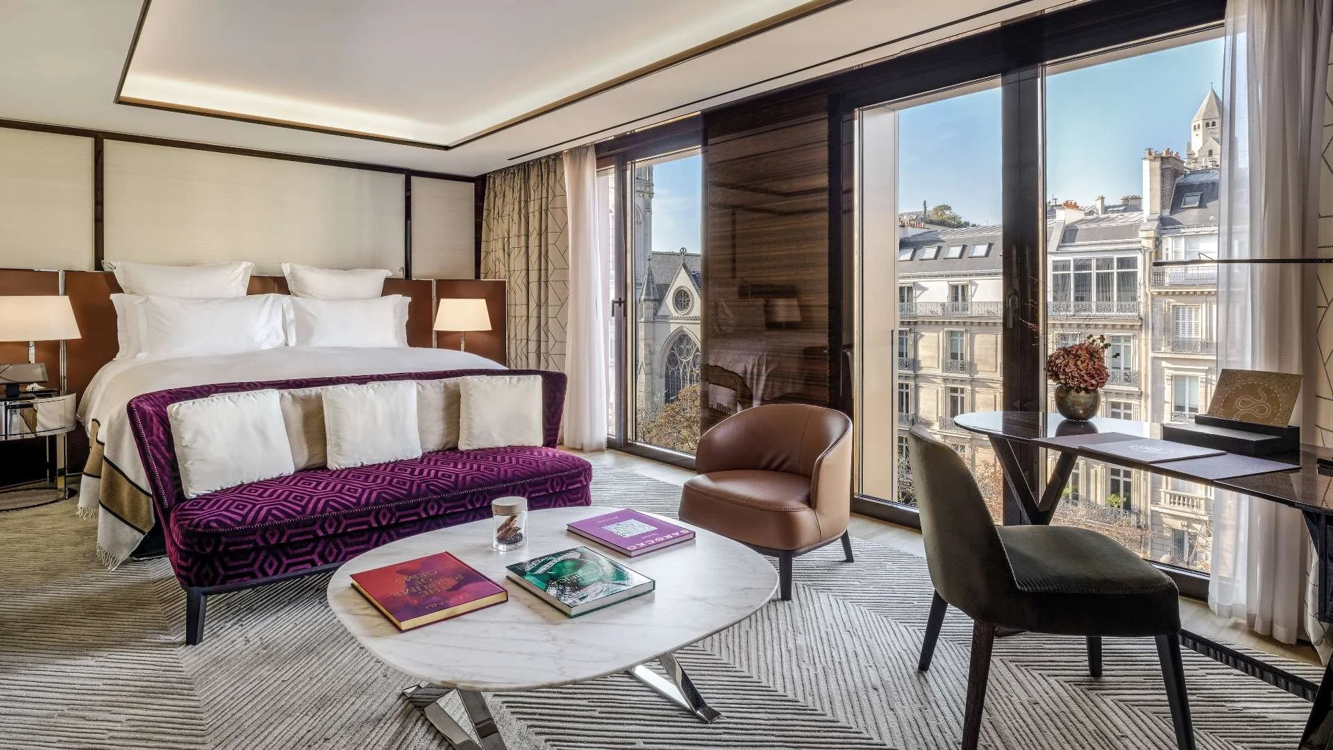 Luxury hotel room with a large bed, purple velvet loveseat, marble coffee table, brown leather chair, black desk with flower vase, and city view through large windows.