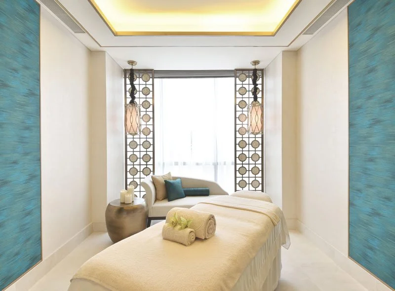 A luxurious spa room with a massage bed, rolled towels, a small sofa with pillows, decorative screens, and soft lighting.