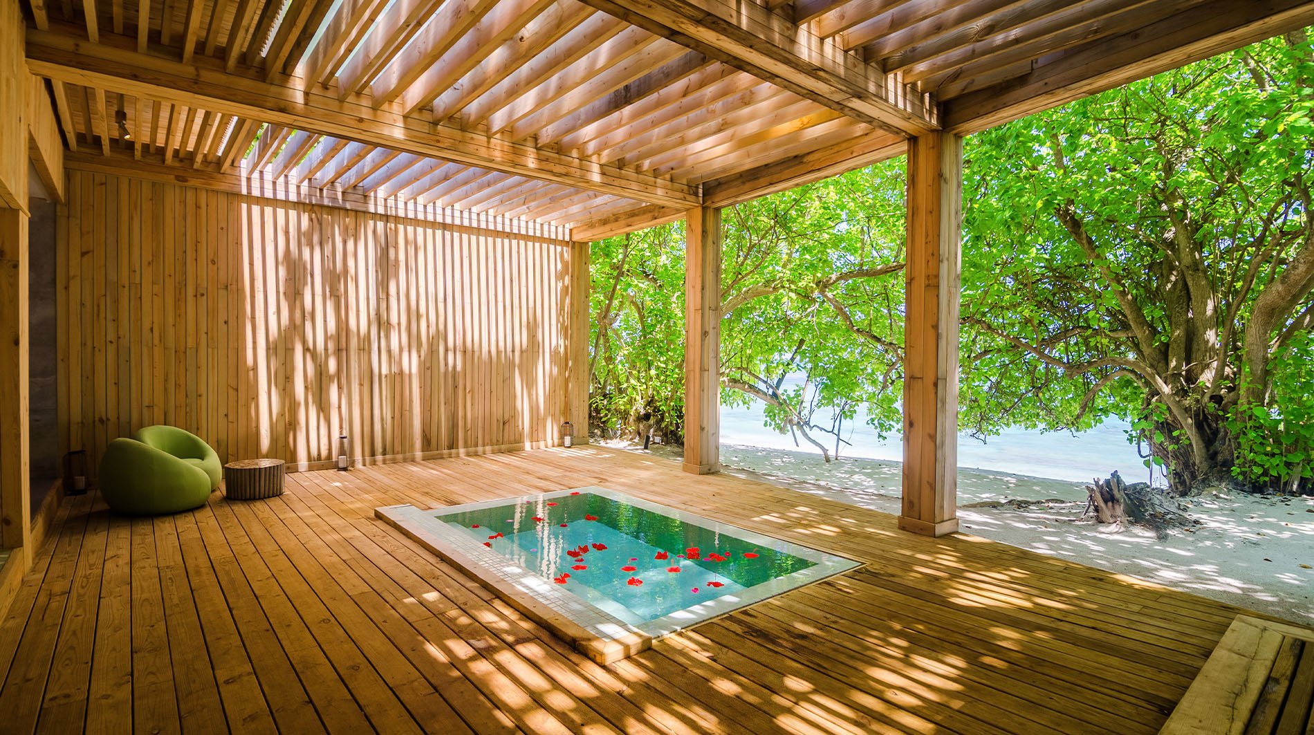 A wooden porch with a small swimming pool, green chairs, and a tree with bright green leaves outside, overlooking the beach.