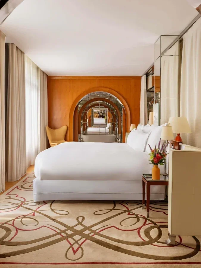A bedroom with a large white bed, side table with a flower vase, yellow armchair, numerous mirrors, and cream curtains.