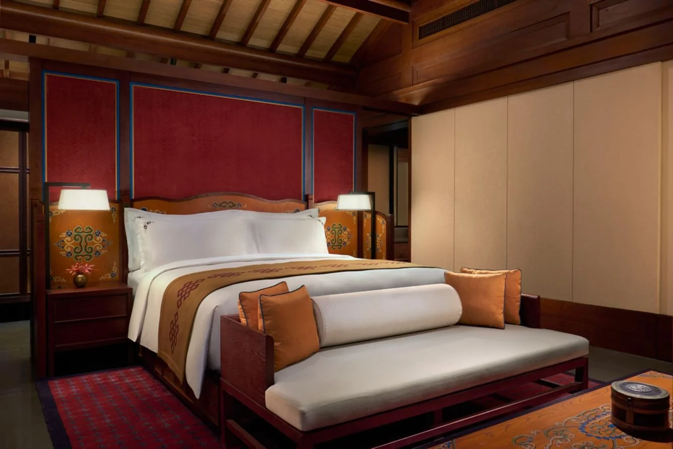 Luxury hotel bedroom with wooden decor, a large bed with white sheets, and a bench at the foot of the bed with cushions.