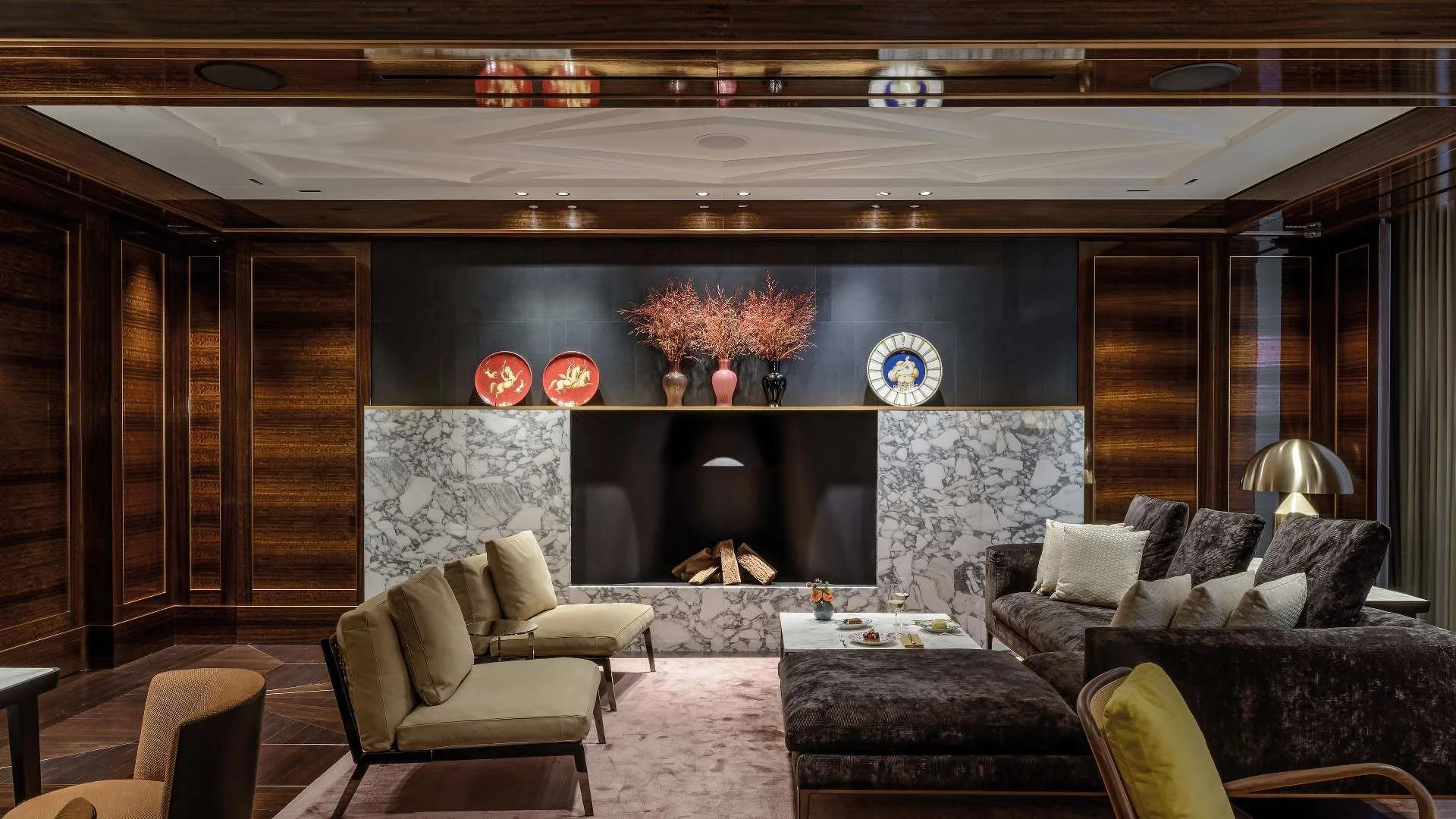 Living room with dark wood paneling, a marble fireplace with logs, beige and dark grey velvet sofas, a marble coffee table with snacks and drinks, decorative vases with pink and red flowers on a shelf above the fireplace, and a gold table lamp.